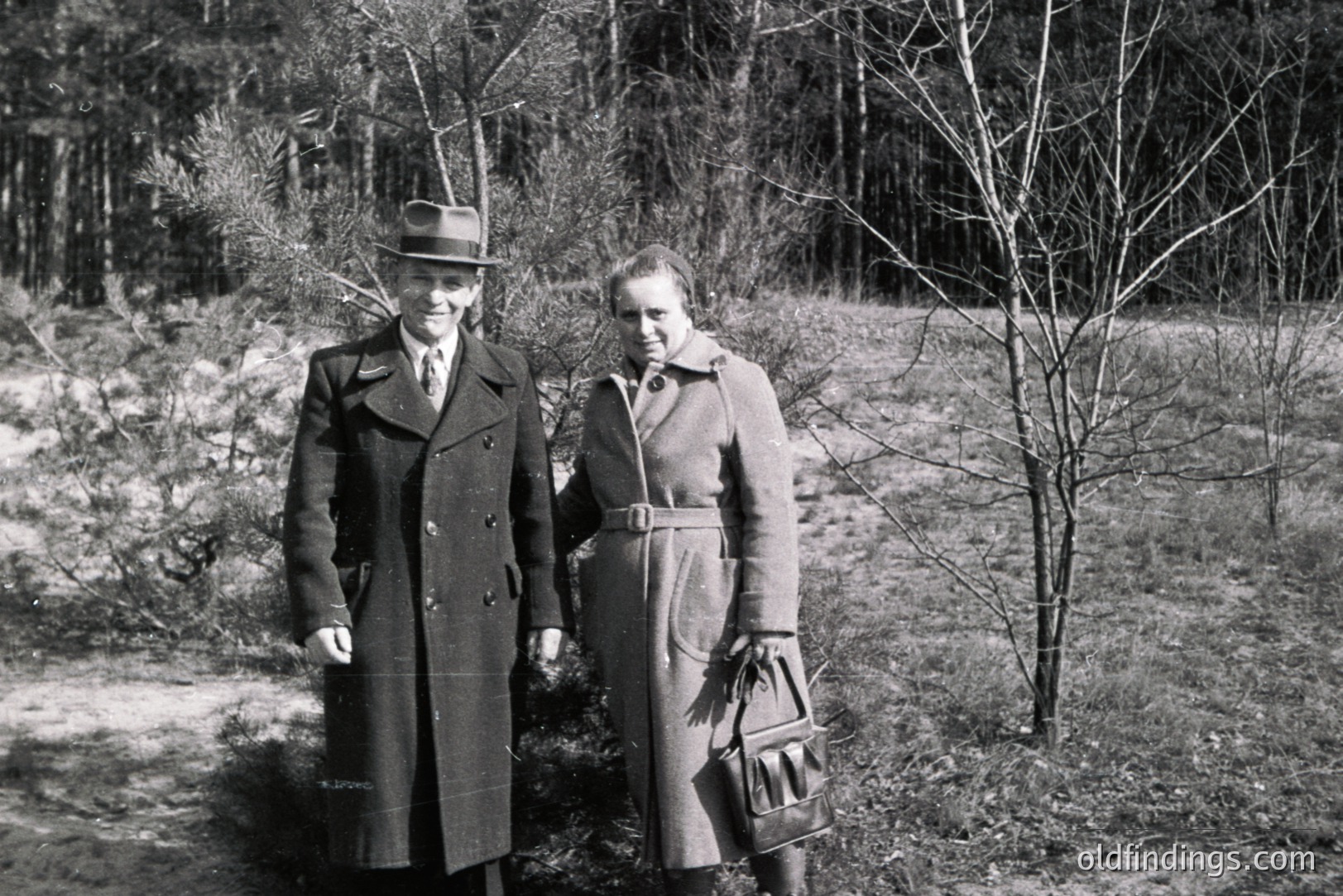 A formally-dressed couple stands outdoors in a sparse, wooded area. The man wears a double-breasted coat, fedora, and tie; the woman, a belted coat and carries a large handbag. Likely 1950s, captured with a distinct mid-century aesthetic. Potential stock photo reference for fashion or period drama.