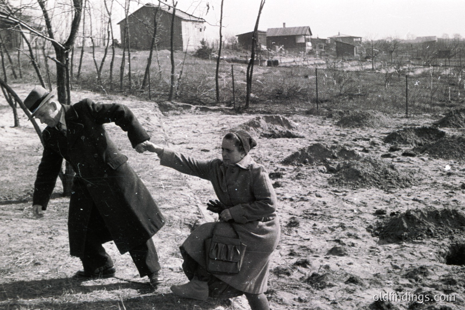 A man in a long coat and hat assists a woman in a coat and gloves, who appears to be struggling on uneven, muddy ground. Likely an outdoor scene, with modest houses visible in the background, suggesting a rural or suburban location. Appears to be mid-20th century.