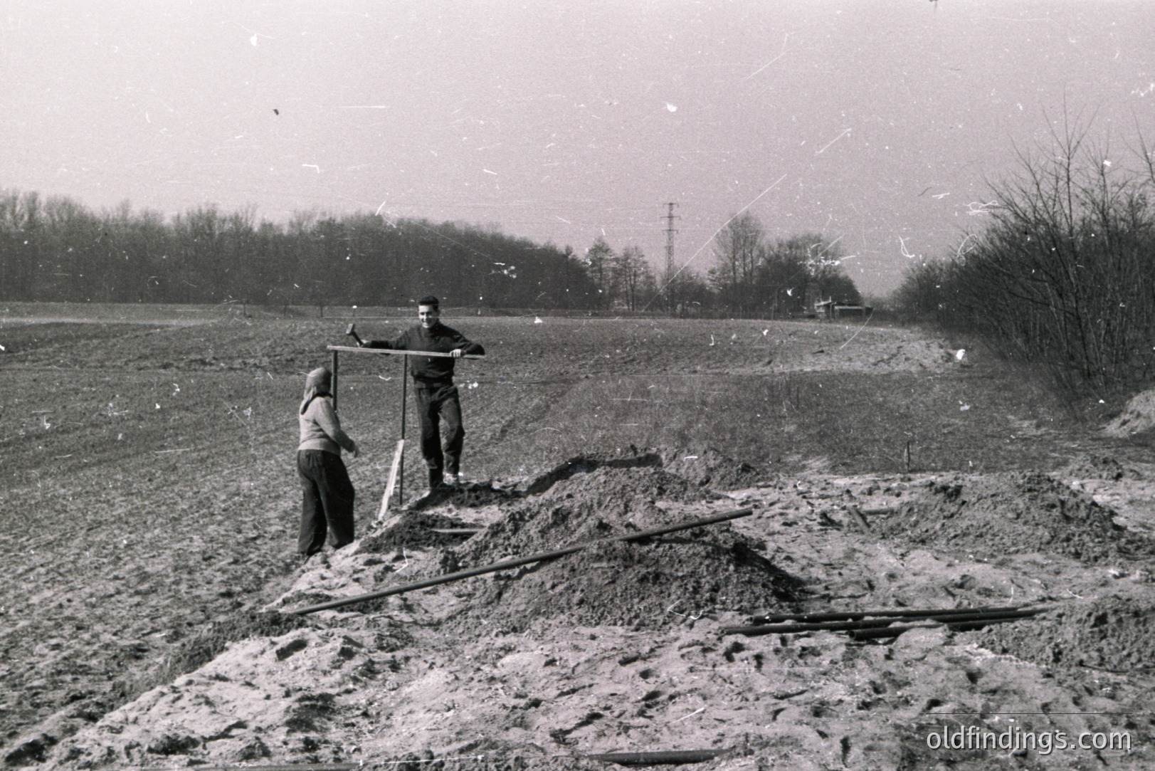 A young man and a child stand near a freshly-dug landscape, likely a construction site or excavation. Earth mounds are prominent, with wooden beams visible. Sparse trees line the background. Appears to be a rural setting, potentially mid-20th century.