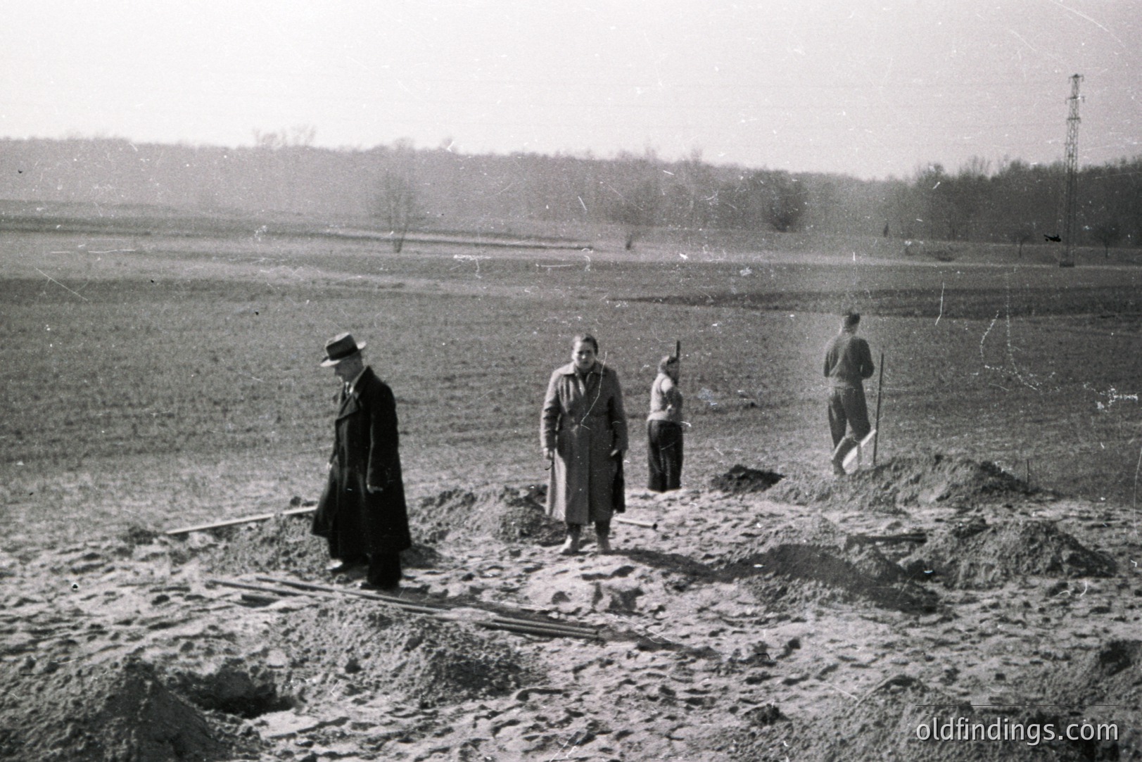 Group of figures observed on a sprawling, excavated earthworks site. Four individuals—likely engineers or supervisors—stand amidst mounds of soil, with tools like rakes and shovels visible. The landscape is flat, bordered by sparse trees. Appears to be a construction or excavation project. Likely 1950s or 60s.