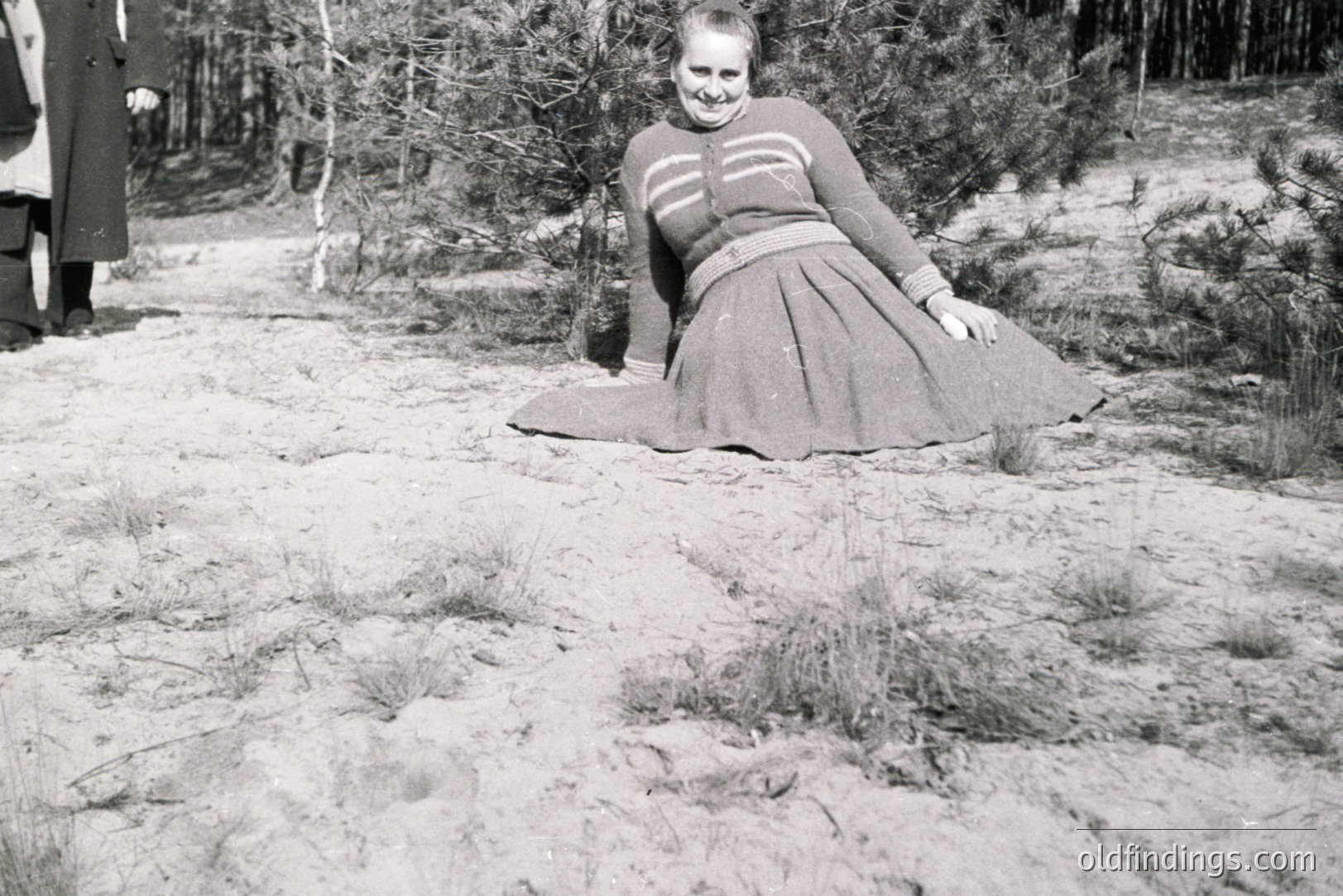 A woman in a textured sweater and full skirt poses in a sandy, grassy landscape, partially obscured by pine trees. A person in a dark coat stands in the background, slightly out of focus. Likely 1950s or 1960s fashion. Potential for design inspiration or vintage aesthetic stock.