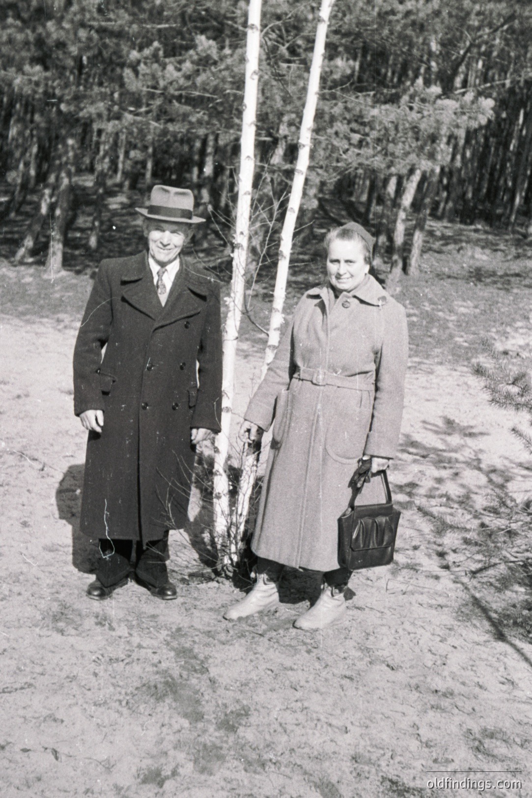 A formally-dressed couple stands near birch trees in a wooded area. The man wears a long overcoat and fedora; the woman, a belted coat and carries a handbag. Likely 1960s or 70s fashion. Landscape suggests a Nordic or Eastern European setting. A simple, timeless portrait.