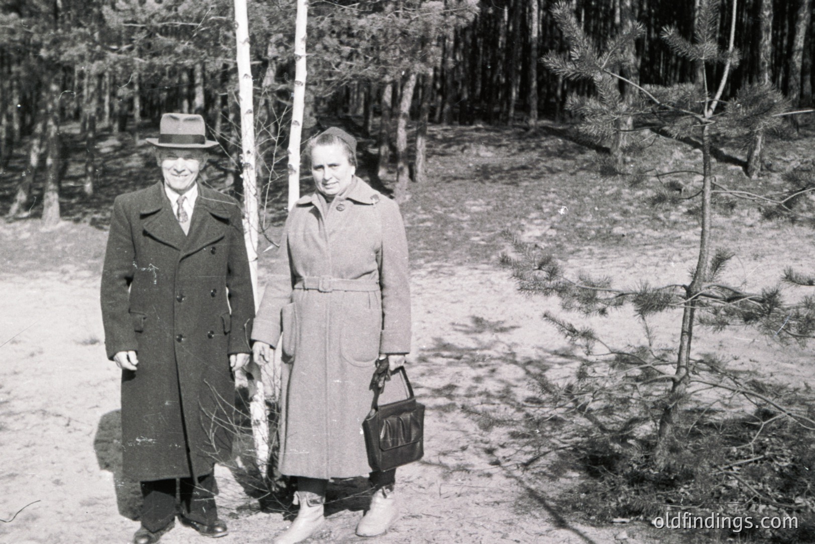 A formally dressed man and woman pose near birch trees in a wooded area. The man wears a double-breasted coat, fedora, and tie; the woman, a belted coat and carries a handbag. Likely a casual portrait, circa 1950s-1970s. Appears to be a personal snapshot, potential stock reference.