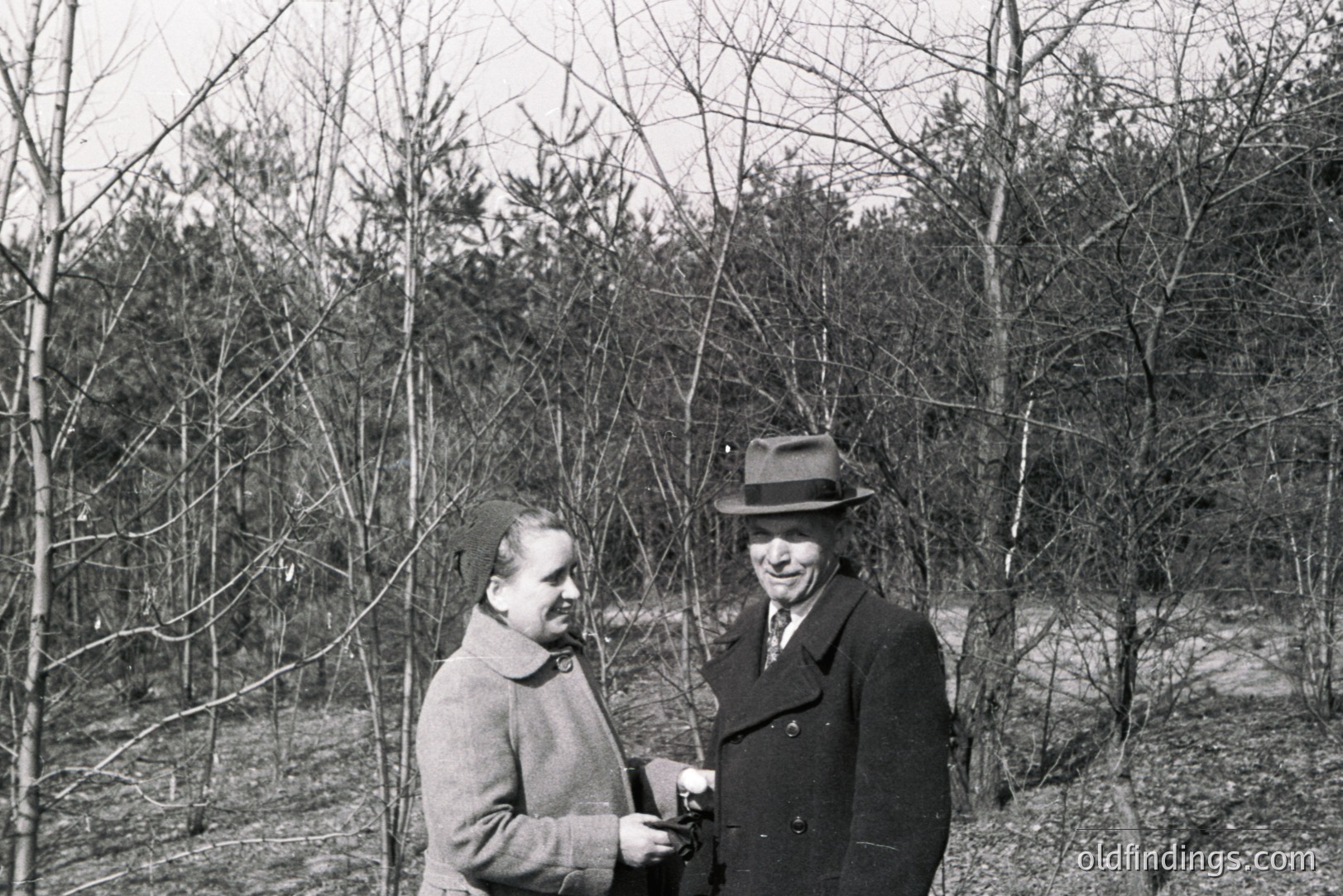 A formally-dressed couple stands amongst bare trees in a wooded area, likely late fall or winter. The woman wears a long coat and scarf; the man sports a peacoat and hat. Appears to be a candid, outdoor portrait, possibly a family snapshot. Likely 1950s or 1960s.