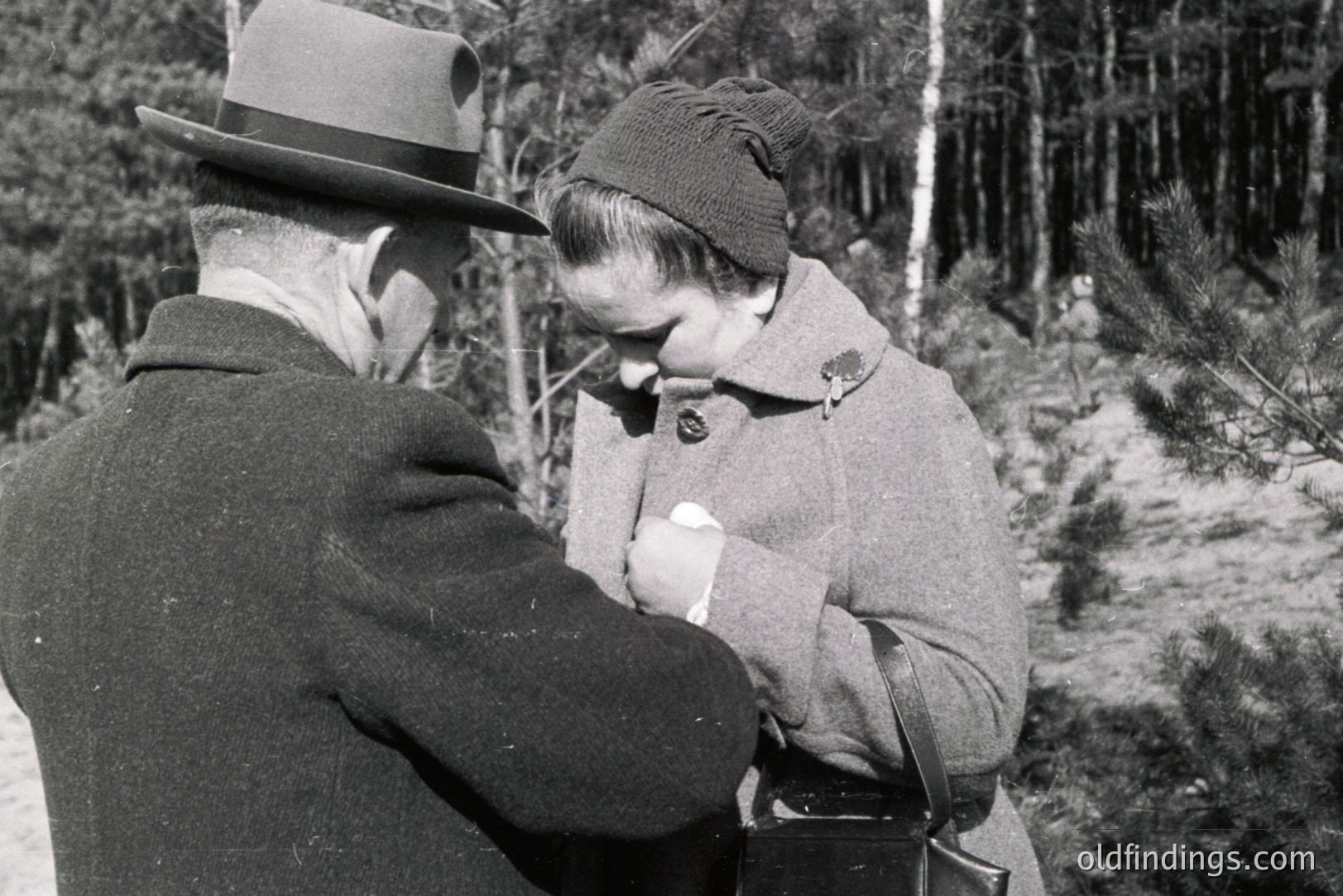 A man in a hat and a woman in a coat appear to share a poignant moment outdoors. The woman holds a tissue to her face, seemingly overcome. Pine trees fill the backdrop, suggesting a wooded area. Likely 1950s-1970s style clothing. A moment of intimacy captured.