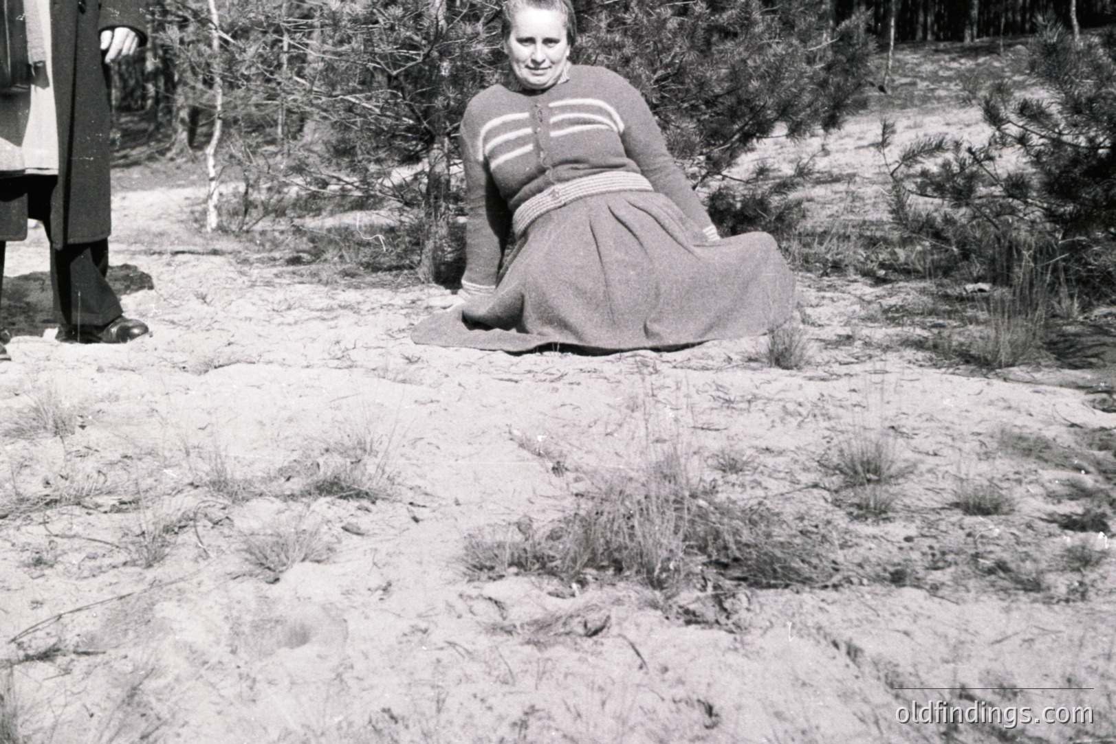 A woman in a full, pleated skirt and sweater sits on a sandy area, partially obscured by pine trees. A man in a dark coat and trousers stands nearby. Likely a candid outdoor portrait, possibly 1950s-1960s. The scene evokes a relaxed, casual moment.