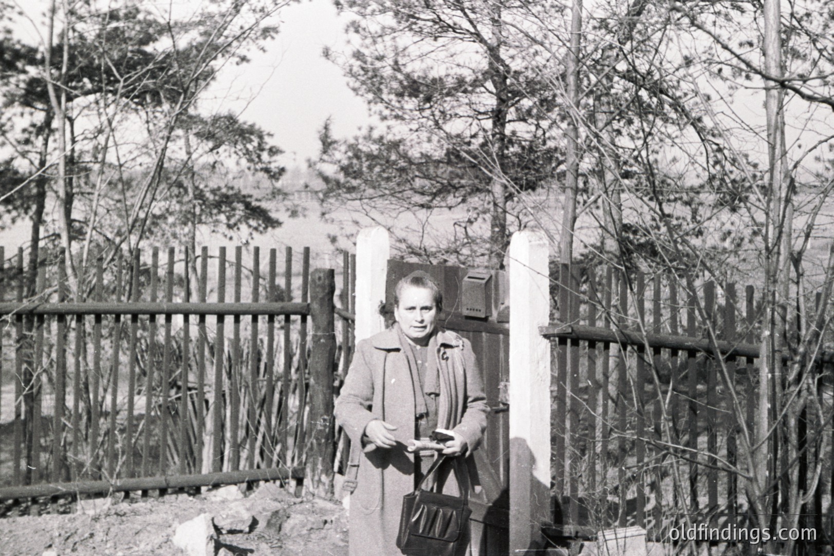 A woman stands at a gate, clutching a small document or card, framed by a rustic wooden fence and a white gatepost. The setting appears to be a rural area with scattered trees. Likely a snapshot from the 1960s or 70s, capturing daily life. Potential stock photo use for nostalgia or travel themes.