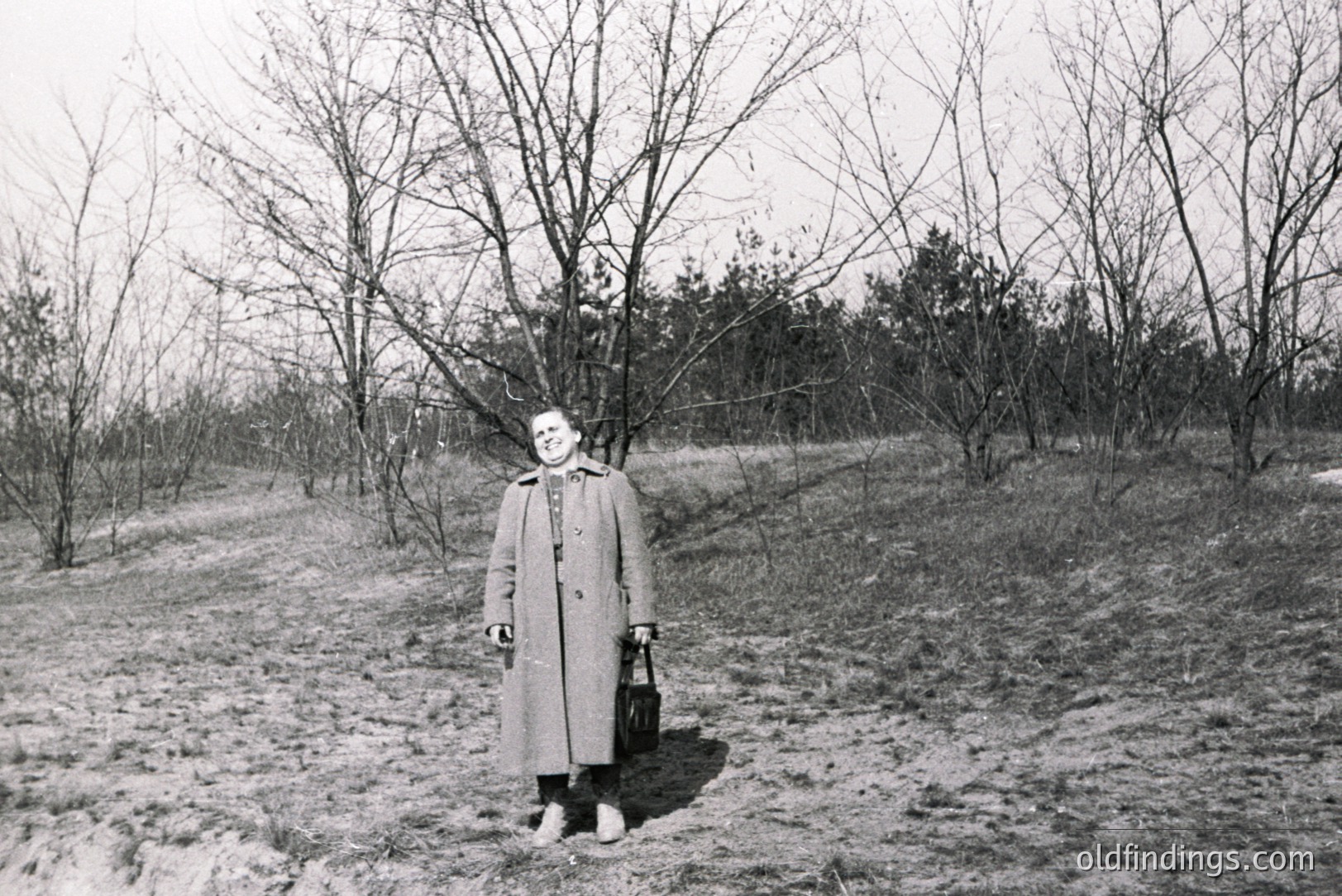 A woman in a long coat stands on a grassy slope, holding a dark bag. Bare trees populate the background, suggesting late winter or early spring. The grayscale image conveys a quiet, contemplative mood. Likely a candid snapshot from the mid-20th century.