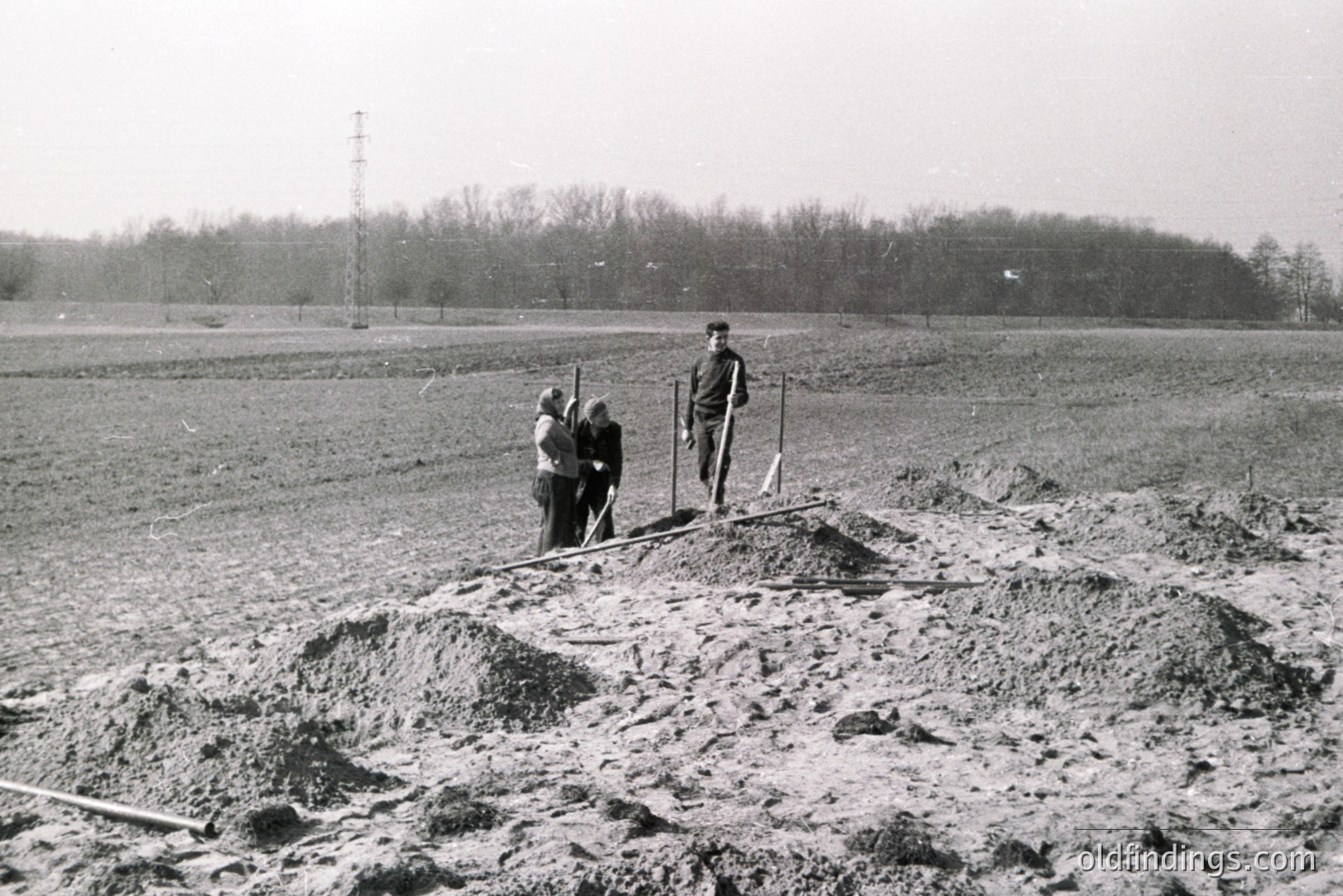 Dusty, monochromatic image depicts a construction site with three figures amidst disturbed earth and piled soil. A man stands centrally, flanked by two figures near the foreground. A communication tower rises in the background. Likely post-war reconstruction or large-scale project.