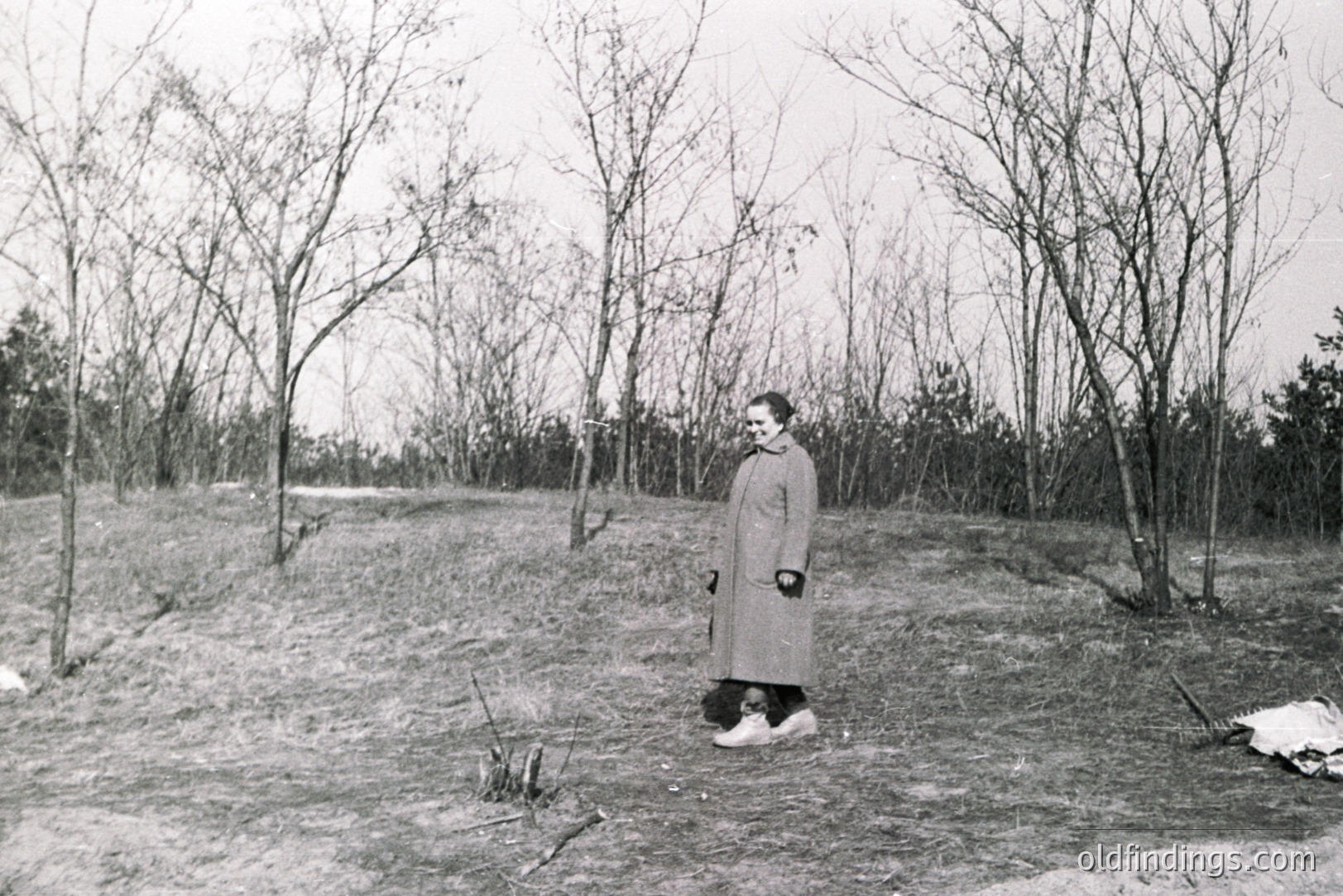 A woman in a long coat stands on a grassy slope, framed by bare trees. Likely a mid-century outdoor portrait, showcasing a stark, natural landscape. The composition suggests a moment of quiet contemplation. Appears to be a documentary or personal snapshot.