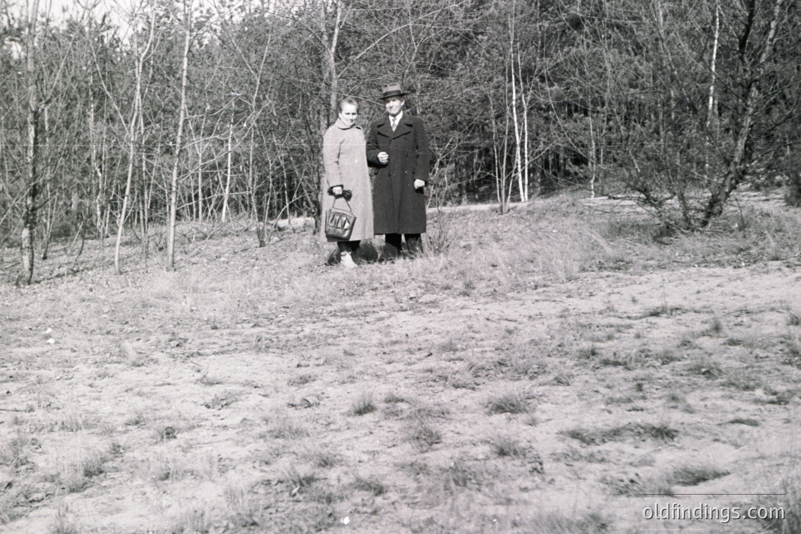 A man and woman stand on a grassy, gently sloping hillside under bare trees. Both are dressed in 1930s-era clothing: the woman in a long coat and the man in a dark overcoat and hat. She carries a small handbag. Possibly a portrait taken during an outing.