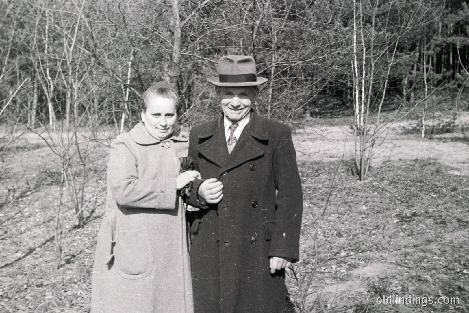 A formally-dressed couple poses outdoors amidst leafless trees, likely in a park setting. The woman wears a long coat with a buttoned collar; the man sports a dark overcoat, fedora, and tie. Appears to be a candid portrait, c. 1950s-1960s. Could be useful for fashion or historical research.