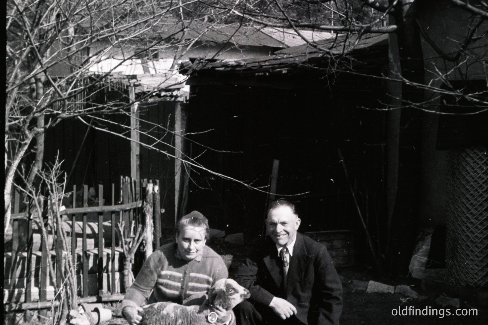 A man and woman, formally dressed, sit with a young lamb in front of a weathered, simple structure, possibly a shed or barn. The scene conveys rural life, likely in a colder climate judging by the clothing. Appears to be a family or posed portrait.