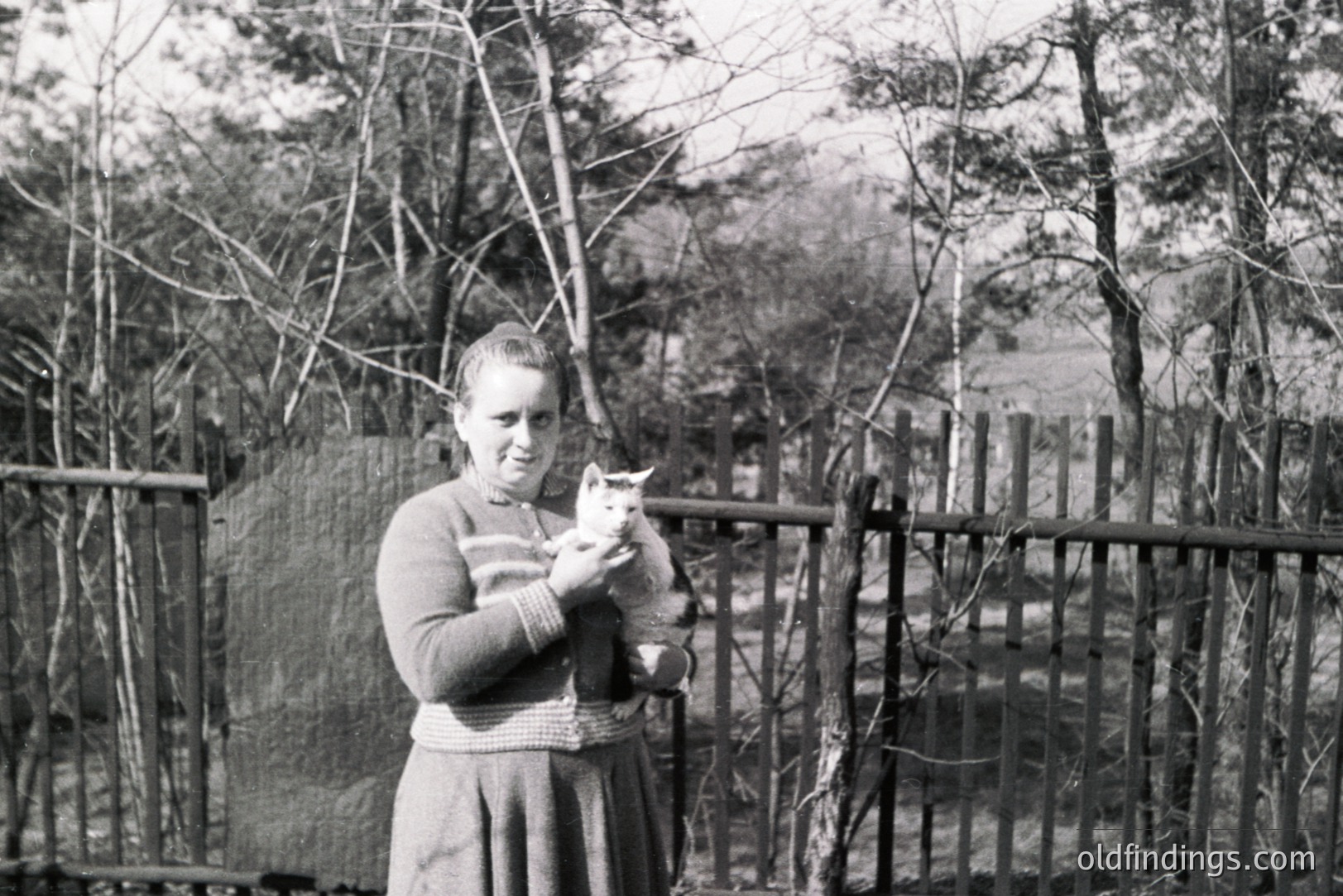 A young woman in a long skirt and sweater holds a kitten outdoors. The scene appears to be in a garden with a split-rail fence and sparse trees. Likely a family snapshot from the mid-20th century. Simple, everyday life captured. Potential stock use for vintage or family themes.