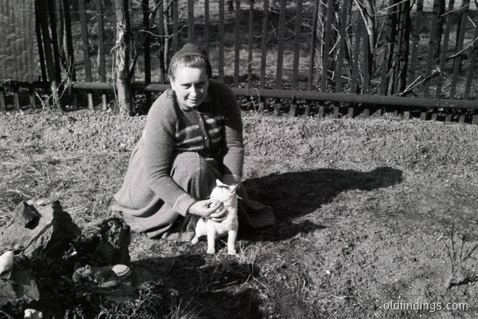 A seated woman cradles a kitten outdoors near a woodpile. She wears a dark, knit sweater and skirt. The backdrop features a weathered wooden fence. Likely mid-20th century rural scene, potentially Eastern Europe. Captures a moment of simple domesticity. Could be valuable for stock or historical context.