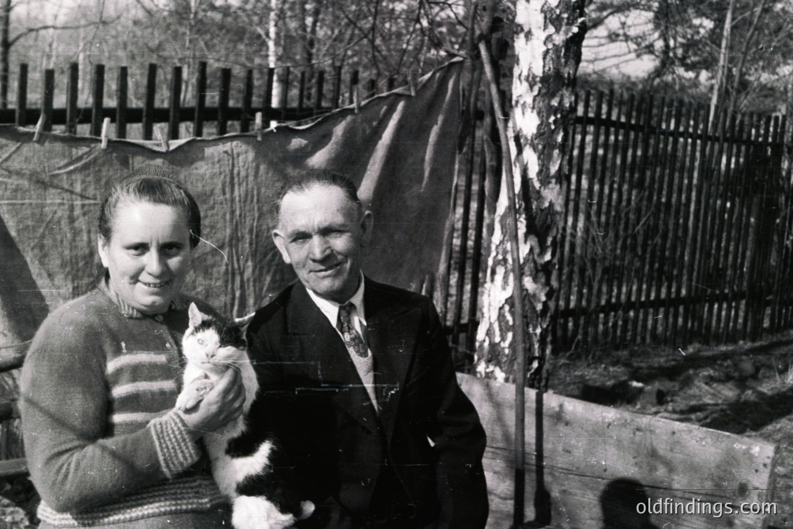 A couple poses in a backyard setting, woman holding a cat. Man wears a dark suit and tie. Rustic wooden fence & tarp backdrop suggest rural or working-class life. Likely a family portrait from the 1950s or 1960s, possibly Eastern European. A glimpse of daily life.
