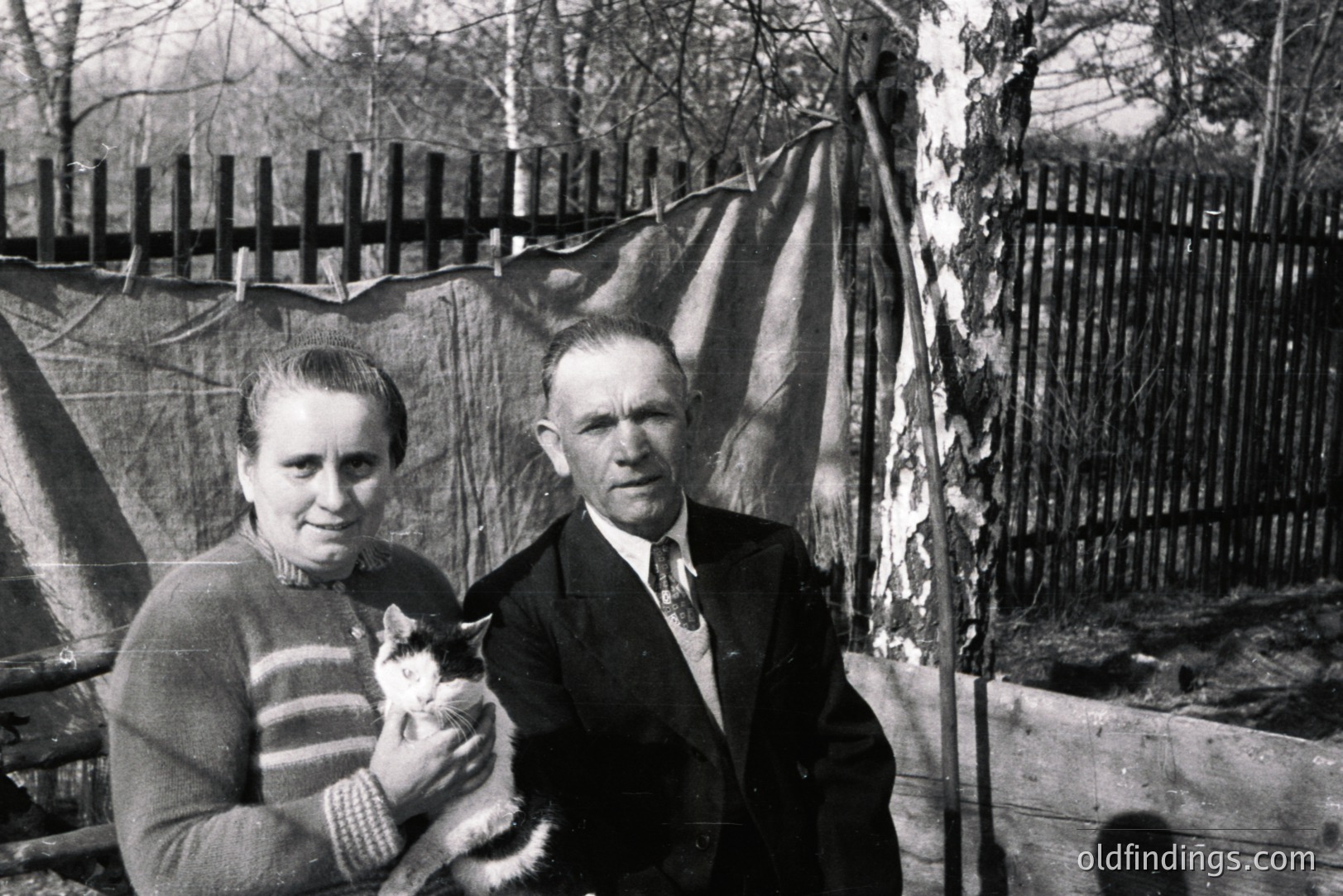 A middle-aged couple stands outdoors, the woman holding a cat. He wears a suit and tie; she, a textured sweater. A canvas tarp and wooden fence form the backdrop, suggesting a rural or transitional setting. Likely a family snapshot, possibly 1950s-1970s.
