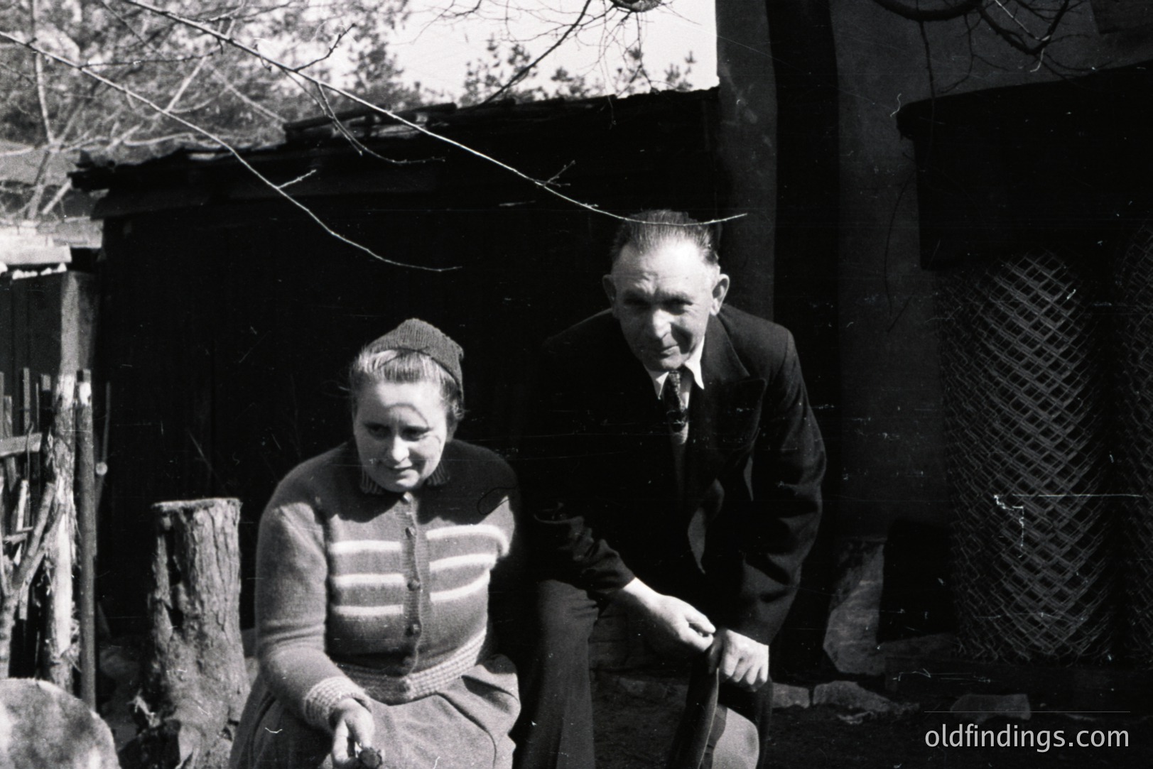 A man and woman sit outside a rustic, whitewashed building with a timbered roof. The man wears a suit and tie; the woman a cardigan and beret. Sparse foliage and weathered wood suggest a rural setting, possibly Eastern Europe. Likely taken in the 1950s or 60s. Potential for historical, genealogical interest.