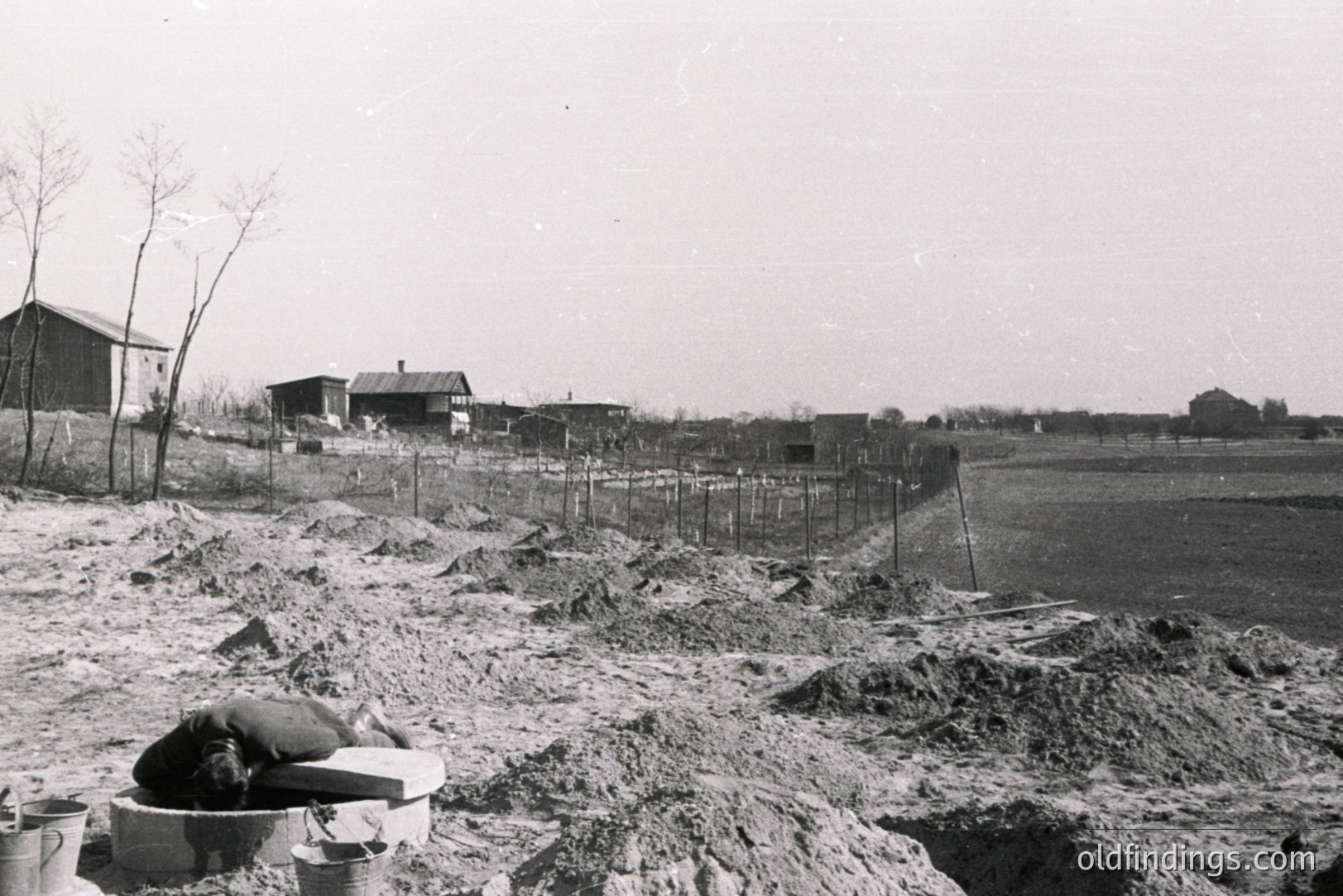 A laborer rests on a makeshift platform amid construction debris and excavated earth. A barbed-wire fence separates the construction site from a sparse rural landscape with scattered farmhouses. Likely a post-war reconstruction or agricultural development project. Appears to be from the mid-20th century.
