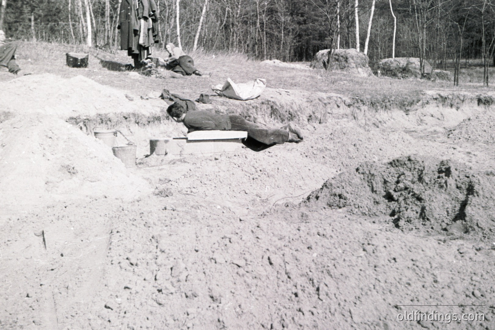 Black and white image documenting a suspected excavation site. A body lies on a wooden board within a newly-dug trench. Several figures are visible in the background, seemingly observing the scene. Likely a forensic or archaeological investigation.
