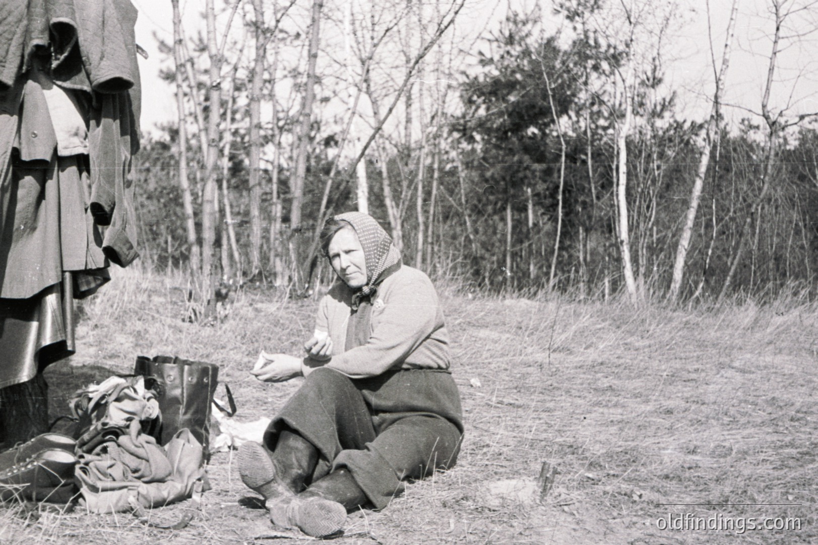A woman in traditional attire sits outdoors amongst scattered belongings and a hanging garment. She wears a headscarf, dark skirt, and appears to be examining a piece of paper. Likely a rural setting, possibly Eastern Europe. Estimated 1930s-1950s. Documenting daily life.