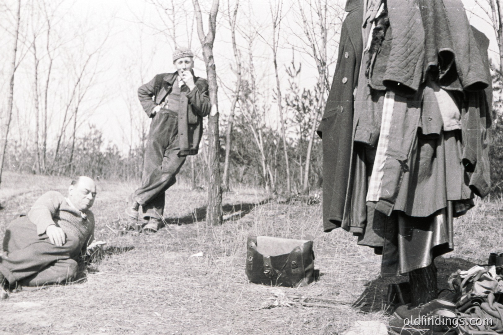 A seated man observes a standing figure amidst a forest setting. Several layers of outerwear and a suitcase lie near a tree. Appears to be an outdoor portrait with a staged or documentary feel. Likely mid-20th century. Potential use for depicting travel, isolation, or fashion studies.