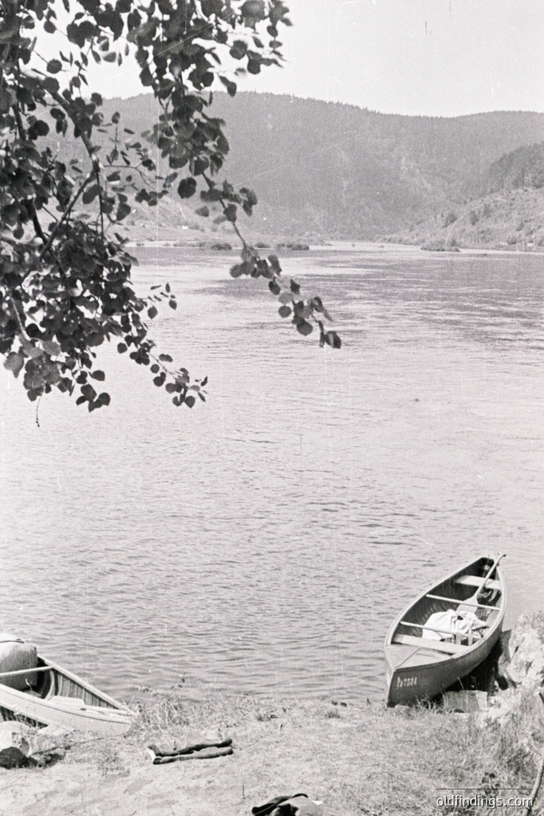 A serene lakescape featuring two small rowboats resting on a grassy shoreline. Gentle slopes rise into the distance, creating a panoramic vista. Foliage drapes the frame's left edge. Likely mid-20th century rural scene, capturing a quiet moment of lakeside leisure.