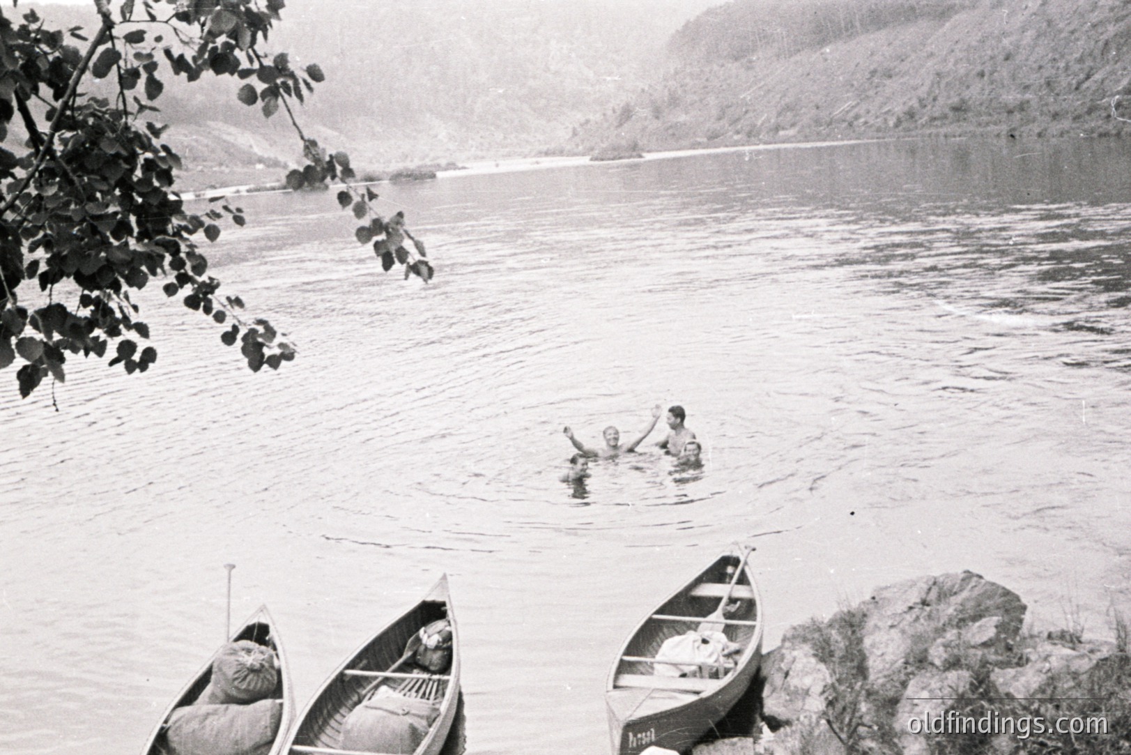 Three wooden canoes are beached near a shoreline with two figures swimming in a wide, placid river framed by steep, forested hills. Likely a recreational scene from the mid-20th century, possibly a summer camp or family outing. Strong potential for stock photography referencing outdoor recreation.
