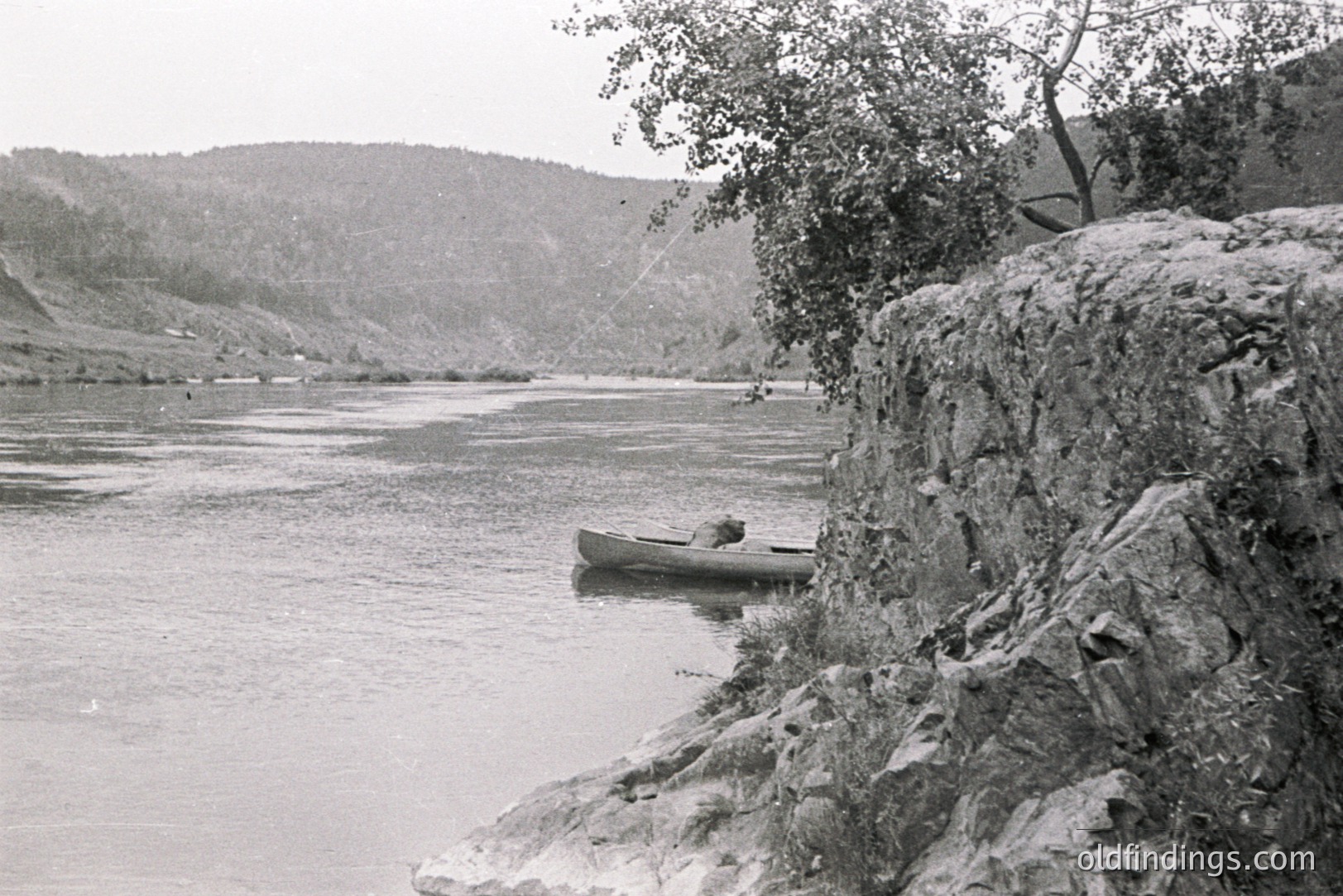 A lone canoe sits on a calm river, with a person visible inside. Rocky shoreline and a tree overhang the water's edge. Rolling hills form a backdrop, suggesting a remote, natural landscape. Likely a scenic travel photo from the mid-20th century.