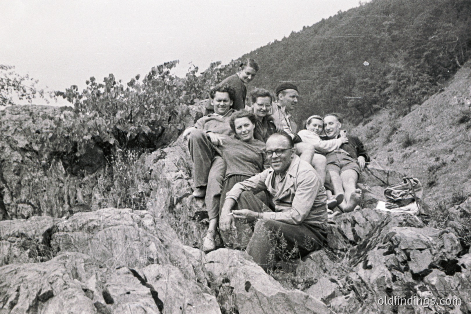 Six individuals pose on a rocky hillside, appearing to be enjoying a leisure outing. The group's attire – casual shirts, trousers, and skirts – suggests a relaxed, informal setting, possibly a holiday. Landscape features indicate an alpine or coastal region. Likely 1950s-1960s. A bag sits near the group.