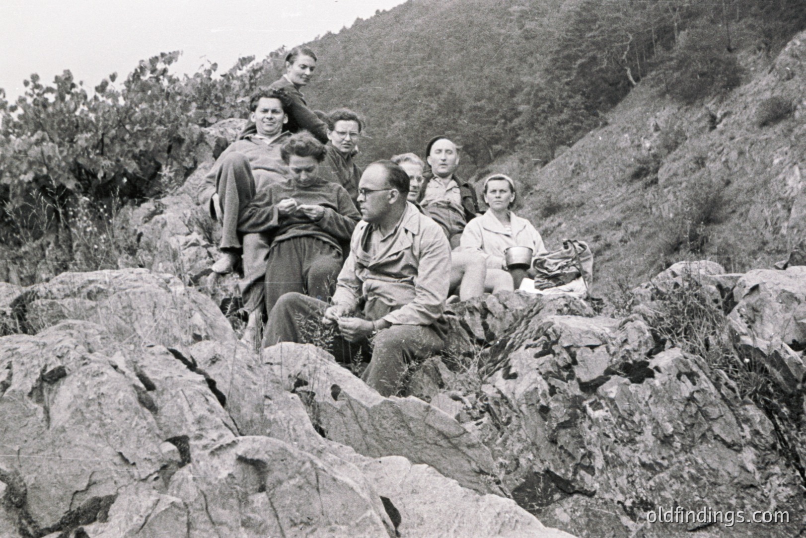 A group of six people pose amidst a rocky, overgrown hillside, likely during a hike or outing. Men wear work shirts & some sport eyeglasses; one woman holds a basket. This appears to be a candid moment, perhaps a vacation snapshot. Likely 1950s-1960s, European origin.