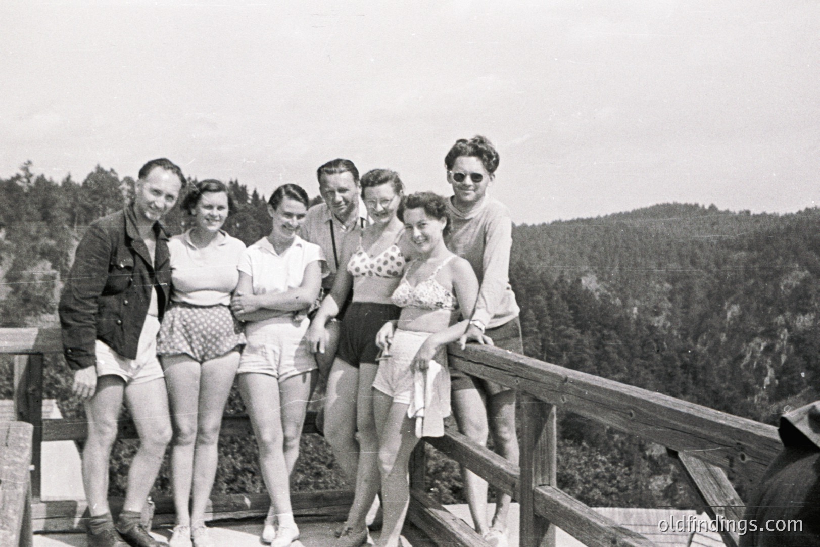 A group of six people pose on a wooden balcony overlooking a forested hillside. Mid-century swimwear and casual wear suggest a vacation setting. Four women wear two-piece swimsuits or shorts, while two men wear casual shorts and a denim jacket.