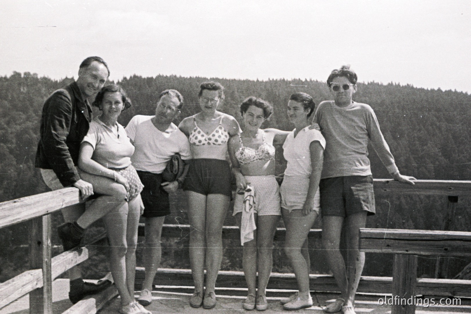 Group portrait, six individuals posing on a balcony overlooking a dense forest. Appears to be a seaside resort or vacation spot, likely 1960s. Fashion indicates summer leisurewear. Balcony's railing and dress styles suggest a mid-century aesthetic. Strong potential for stock photography & design reference.