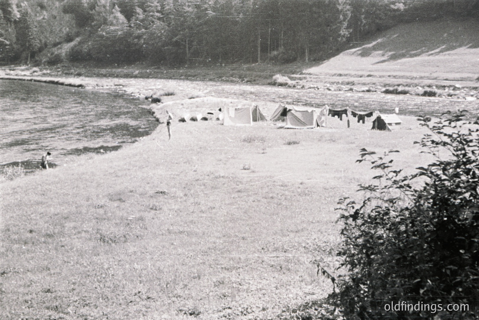 A riverside campsite features several canvas tents arranged along a grassy bank. A lone figure stands near the shoreline. Dense trees line the background, suggesting a secluded, natural setting. Likely a recreational or scouting group gathering, circa 1950s-1970s.