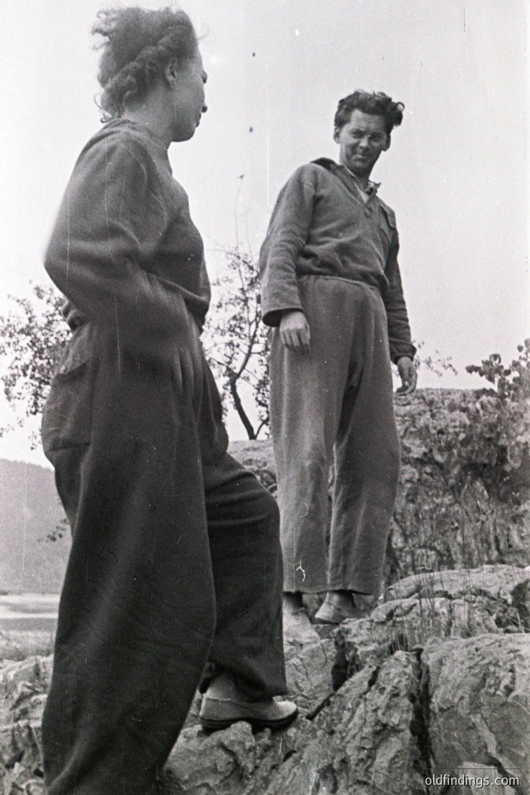 Two figures are captured on a rocky, coastal ledge. A woman in wide-legged trousers and a loose-fitting top stands observing the view, while a man in workwear gazes in the same direction. Likely a candid moment, possibly depicting a rural or working-class setting. Estimated 1950s-1970s timeframe.