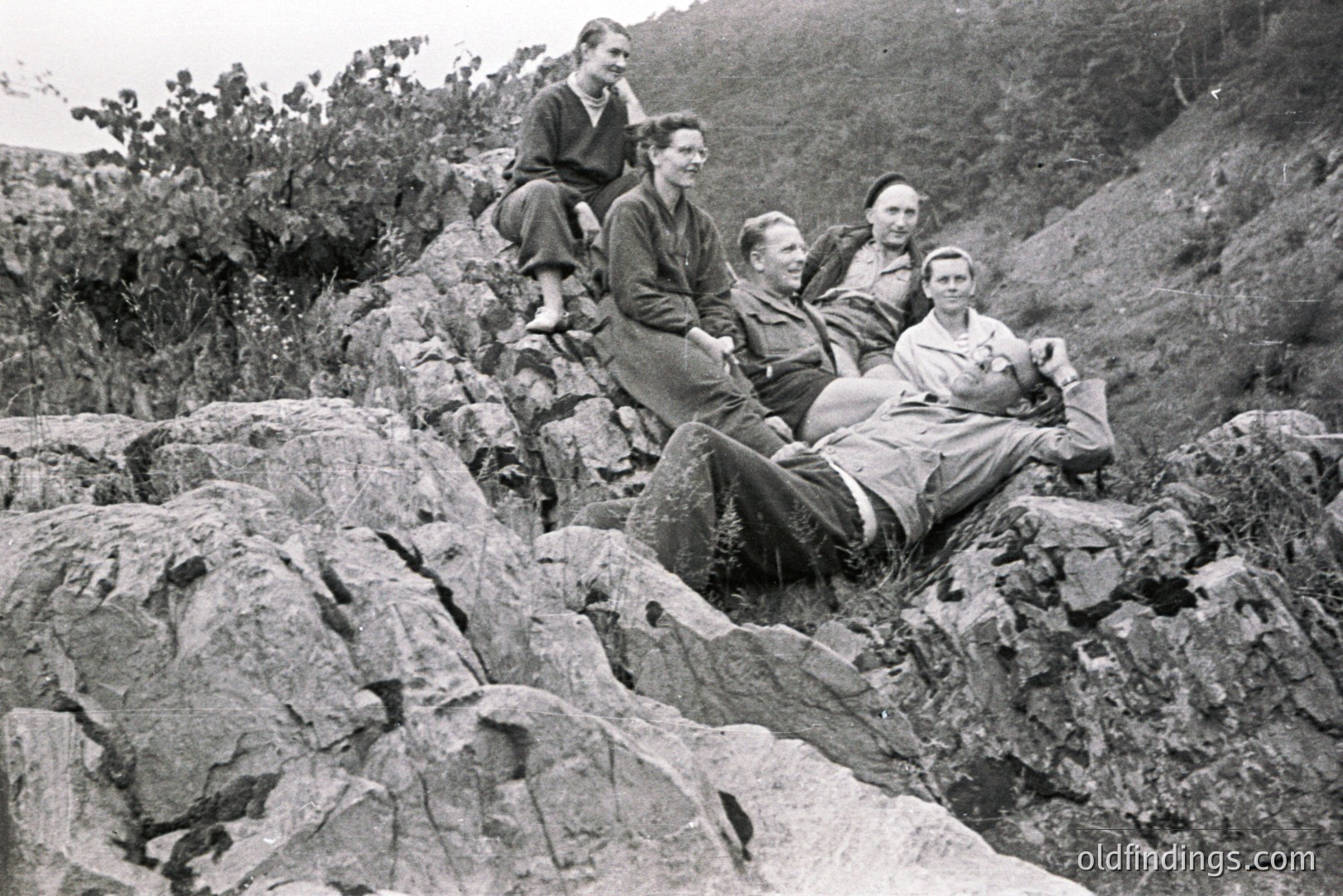 Five individuals casually posed on a rocky hillside; several are seated or reclined on the stone. Clothing suggests a mid-century, possibly 1950s or 60s style. A scrubby landscape rises behind the group. Likely an outdoor leisure scene.