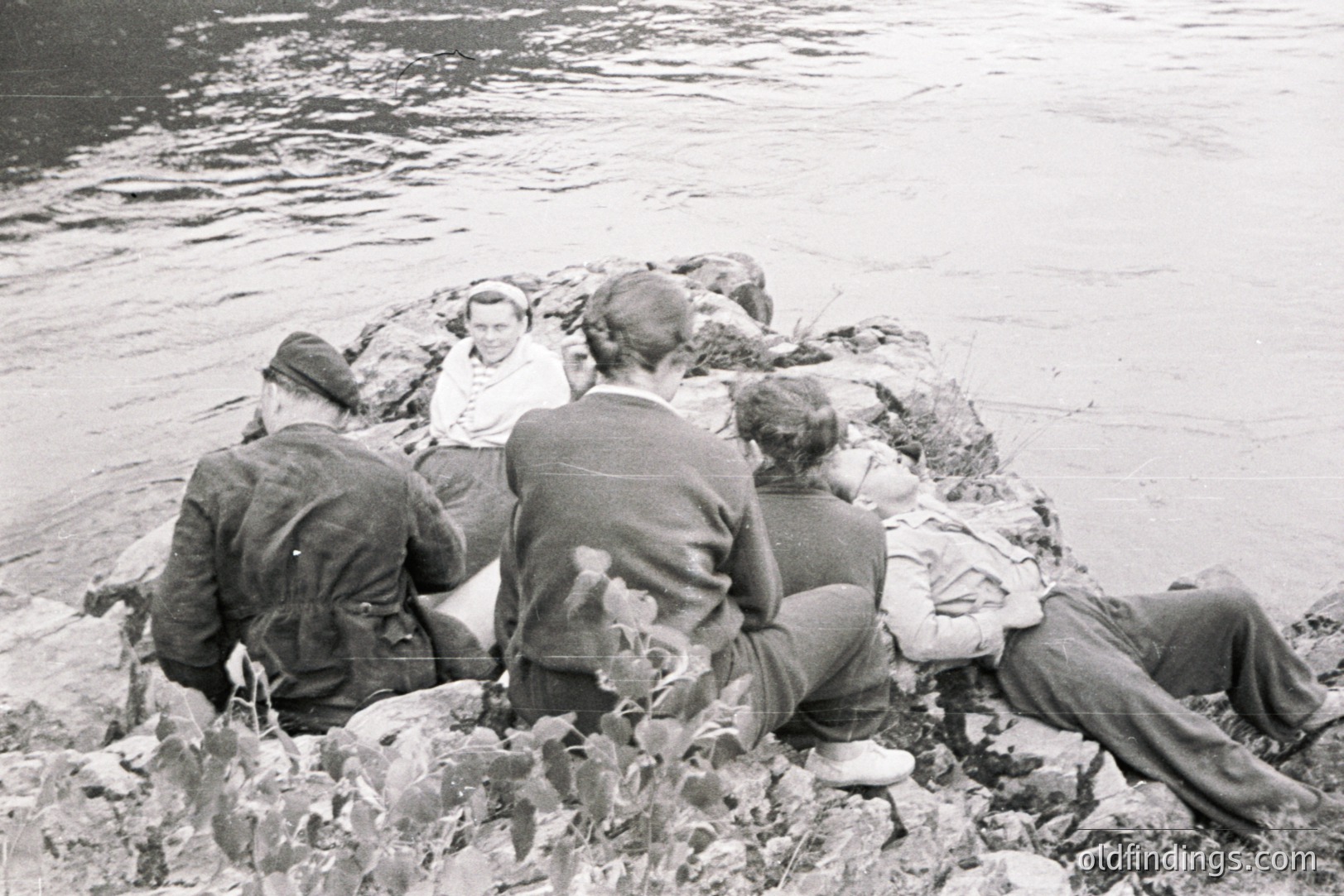 Four people, partially obscured, relax on a rocky outcrop beside a body of water. Three wear jackets and trousers; one wears a dress and headscarf. The scene suggests leisure time, possibly a picnic or outing. Appears to be a candid snapshot, likely from the 1950s or 60s.