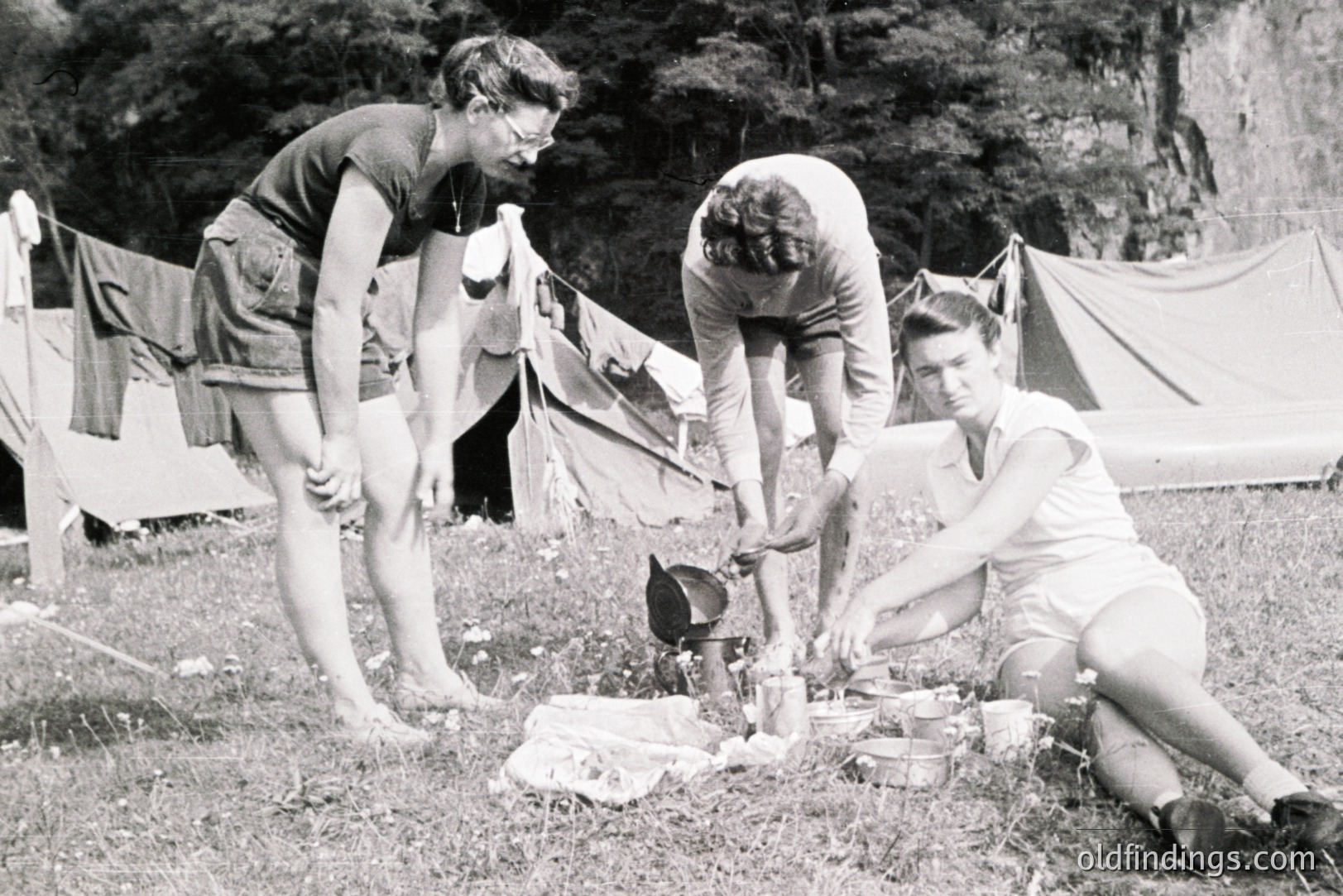 Three young people prepare food outdoors near a tent, seemingly on a camping trip. One is barefoot, another is tending to a small stove and cookware. Clothing is casual, suggesting a relaxed atmosphere. Likely 1950s-1960s, North America.