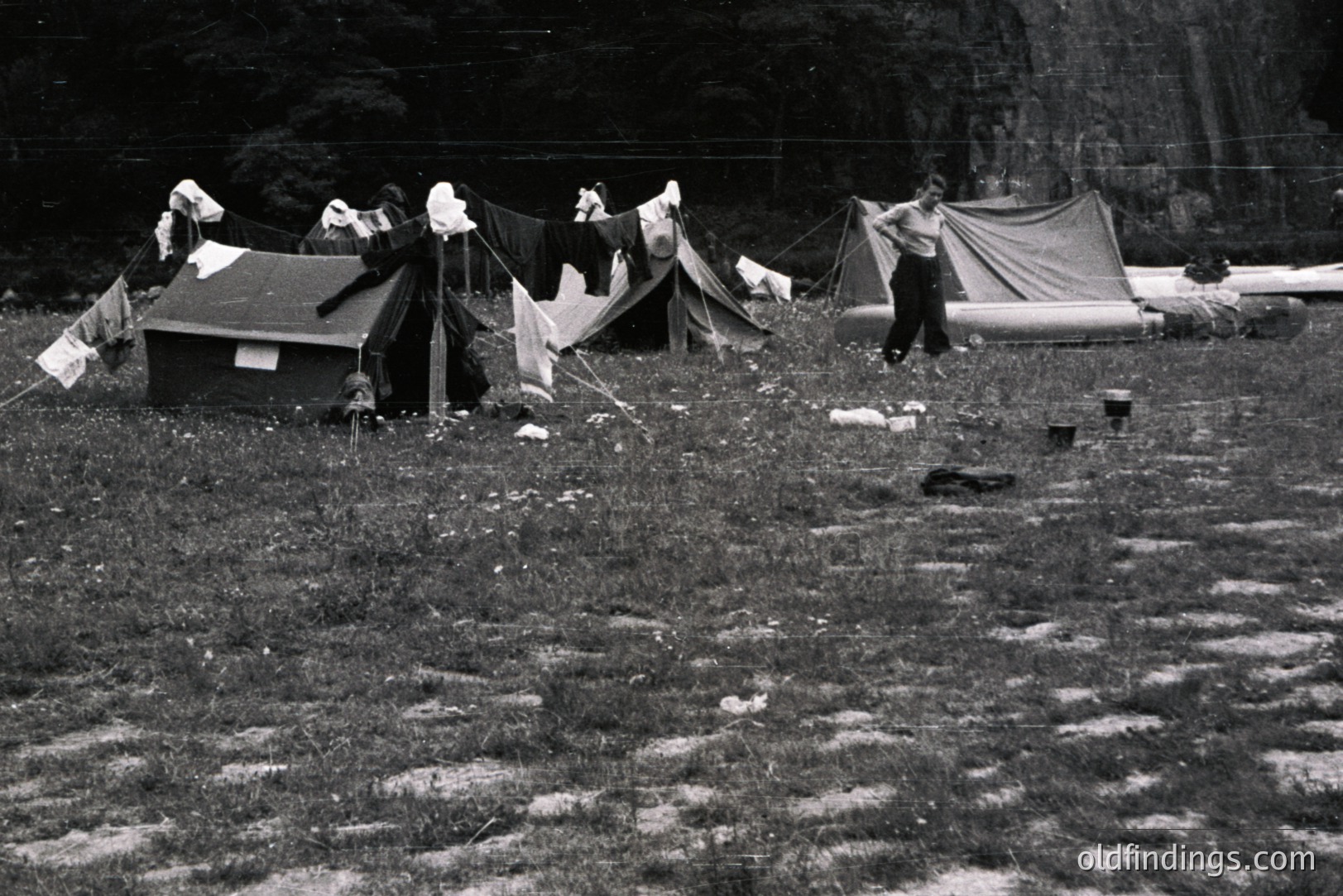 A grassy field houses several military-style tents, with laundry lines strung between them. A soldier stands near a tent, seemingly observing. Evidence of camping gear and canoes suggests a recreational or training activity. Likely post-WWII era, possibly 1950s.