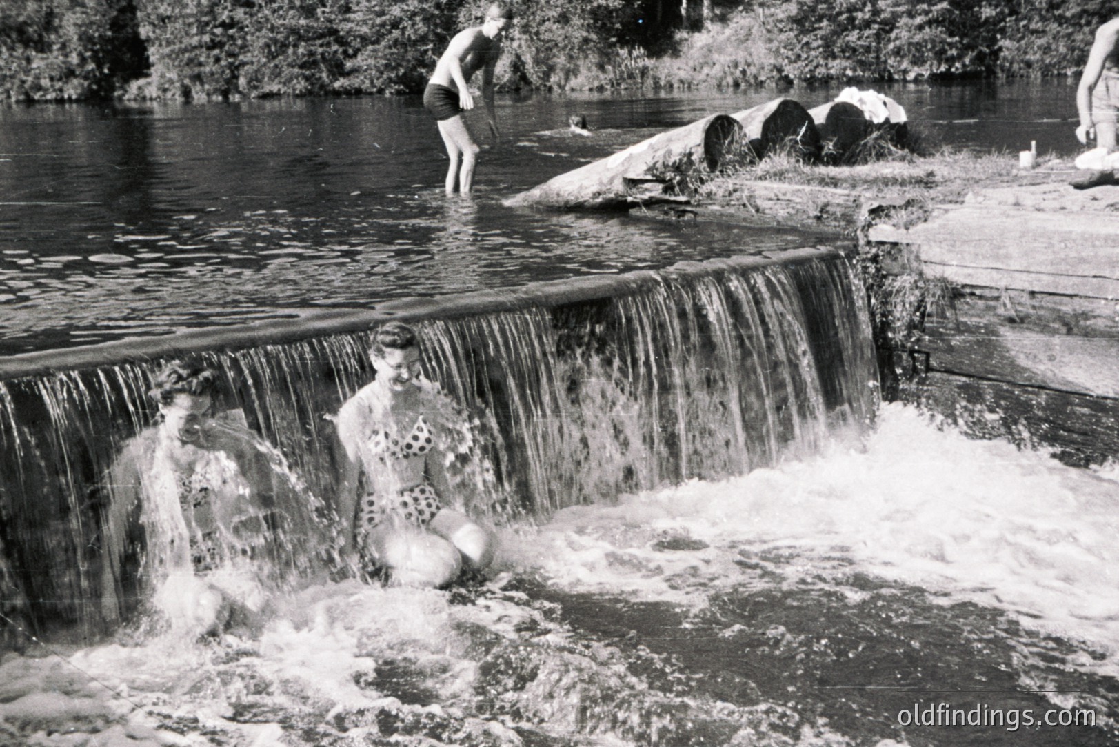 Two women in swimwear pose near a small waterfall, enjoying a riverside recreation area. A young man is jumping into the water behind them. The scene captures leisure activities likely during the 1950s or 1960s. Rural setting with visible foliage and logs. Documentary-style image with potential historical research value.