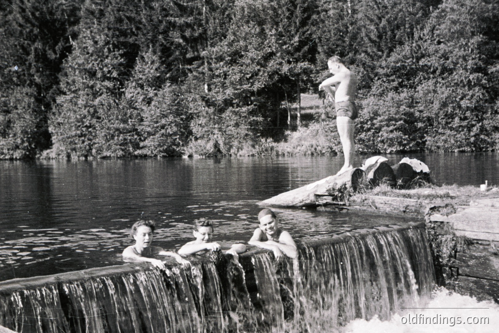 Three young men enjoying a summer swim at a small dam/waterfall. Two are partially submerged, seated atop the concrete structure. A third stands poised on a rocky outcrop in the background. Likely a recreational area, possibly a resort. Appears to be a candid snapshot, circa 1960s.