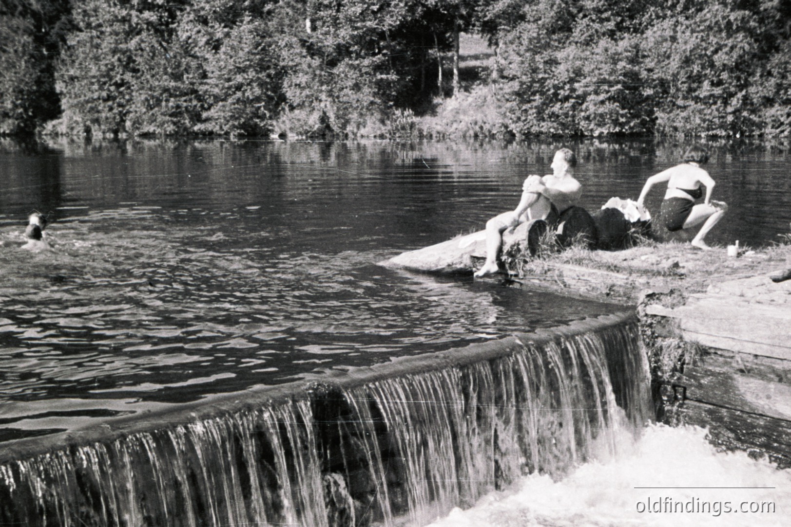 Three young people relax on a stone ledge beside a small waterfall and reservoir. Two are reclining, one appears to be swimming in the water. Likely a recreational area, possibly a dam structure. Black and white photograph, evocative of the 1950s-1970s. Potential for stock or design use.