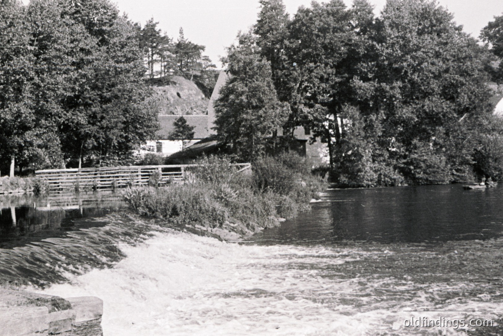 A grayscale shot features a waterway cascading over a man-made stone structure, surrounded by dense foliage & trees. A small building sits atop an earthen embankment in the background. Likely a recreational area or park, possibly 1950s-1970s. A rustic bridge spans the water.