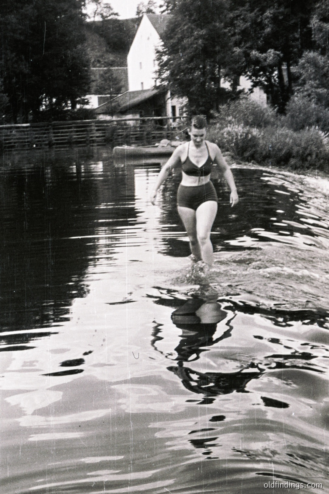 A young woman in a high-waisted, one-piece swimsuit wades through shallow water. A small house and boathouse are visible in the background. The image exhibits clear signs of aging, including scratches and dust. Likely a mid-century candid moment of leisure.