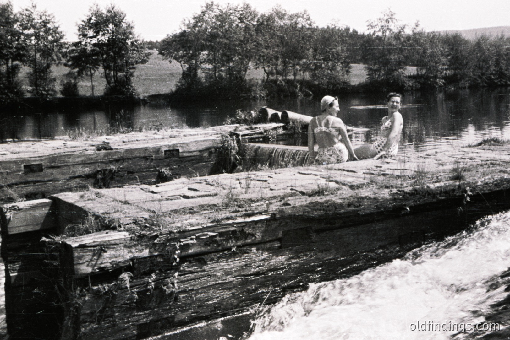 Two young women in swimwear pose on a weathered, decaying wooden structure over a river rapids. The scene captures a playful, casual moment, likely a leisure activity. The landscape features dense foliage and a hillside backdrop. Appears to be a vintage, possibly 1950s-1960s, snapshot.