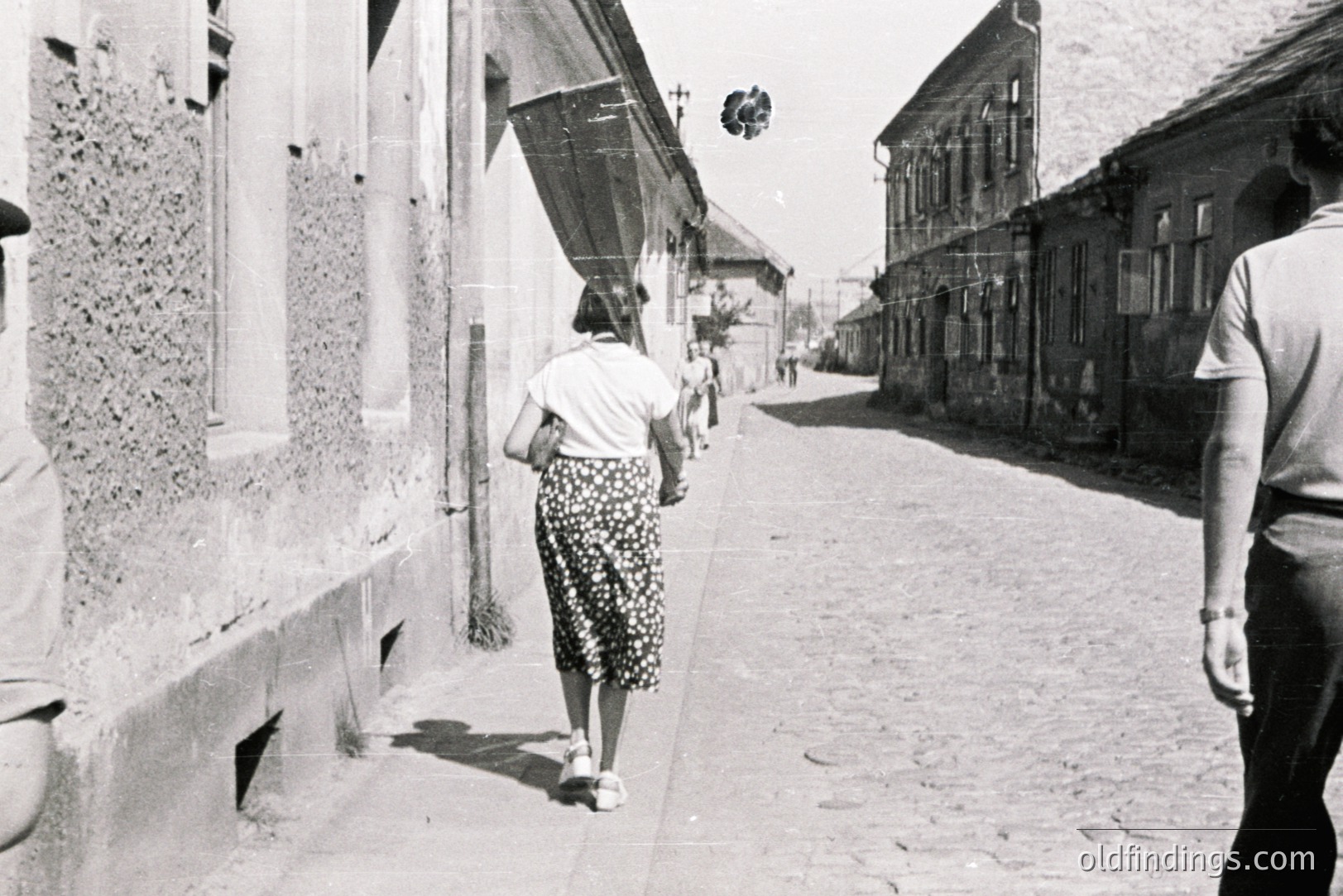 A woman in a patterned dress walks away along a cobbled street lined with aged, weathered buildings. A second figure follows behind. Buildings feature shuttered windows & textured facades. Likely Eastern European, possibly 1960s-70s. A palpable sense of quietude & time.