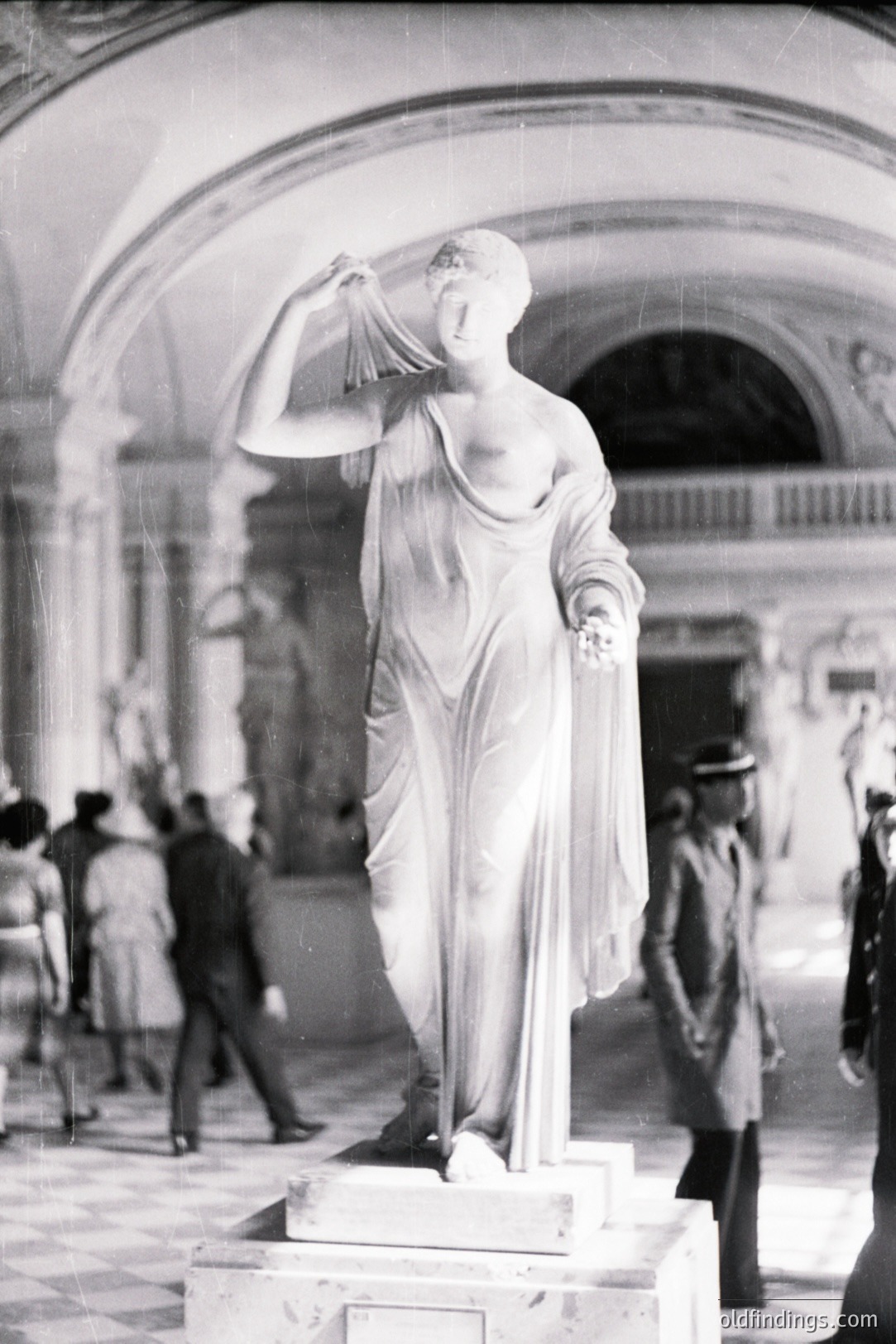 Dramatic black & white photo: a marble statue of a draped female figure stands within an ornate, classical-style interior. Visible are onlookers & uniformed guards. Likely a museum or gallery setting. Appears to be from the mid-20th century. A valuable reference for architectural design & classical art studies.