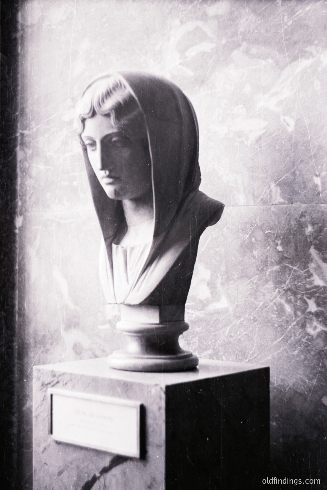 Classical bust of a woman draped in a flowing veil, displayed on a stone pedestal. The marble sculpture exhibits detailed facial features and drapery folds. The background features a textured wall, possibly marble, adding depth. Likely a museum or gallery setting.