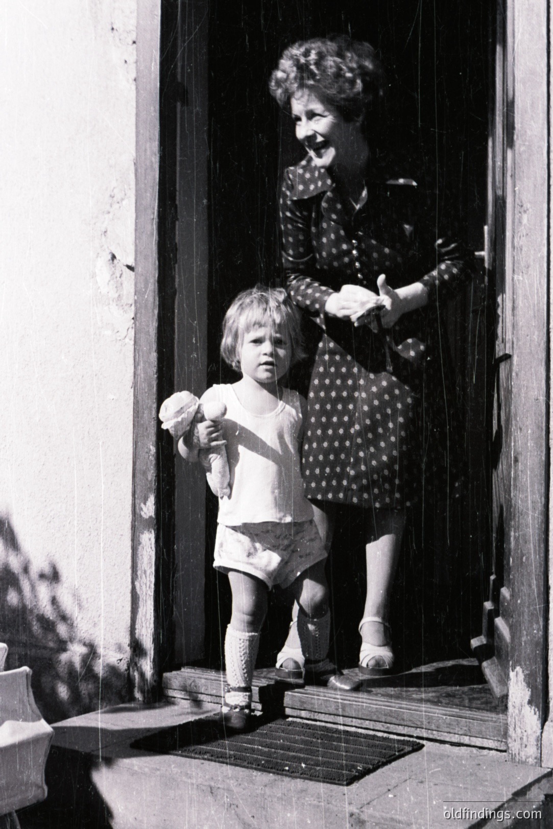 A young child stands at a doorstep, clutching a small toy, while an adult woman, dressed in a patterned dress and sandals, stands partially visible in the doorway. The scene appears domestic, likely a family moment. Architectural details suggest a European setting. Likely 1960s-1970s. Could be valuable for nostalgic stock imagery or historical reference.