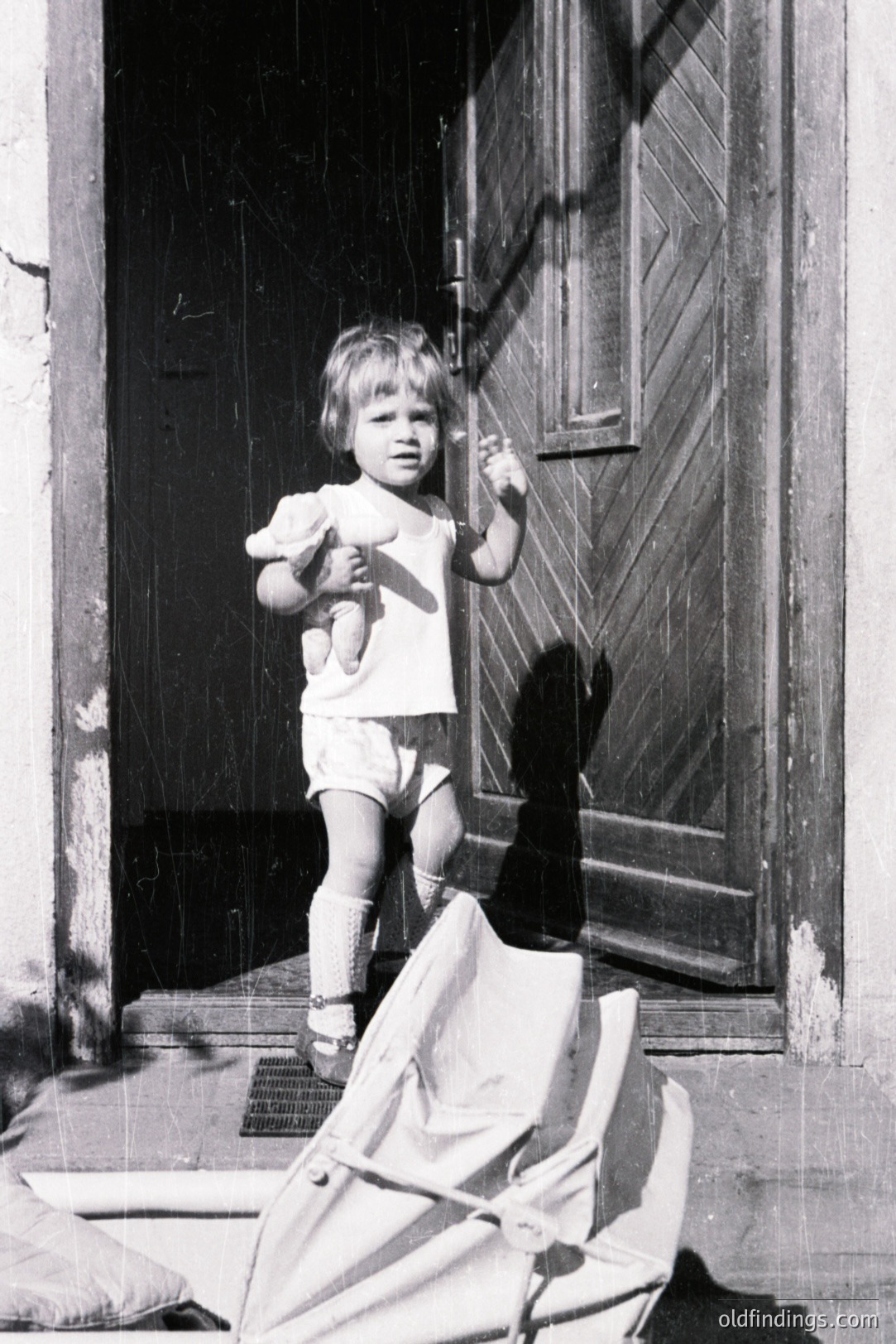 A young child, likely a toddler, stands on a doorstep clutching a teddy bear. Visible in a simple cotton dress and short pants. The aged, weathered wood door and visible shadow suggest an older building. Photograph likely from the 1960s or 1970s, typical for its grainy texture. Strong architectural and documentary value.